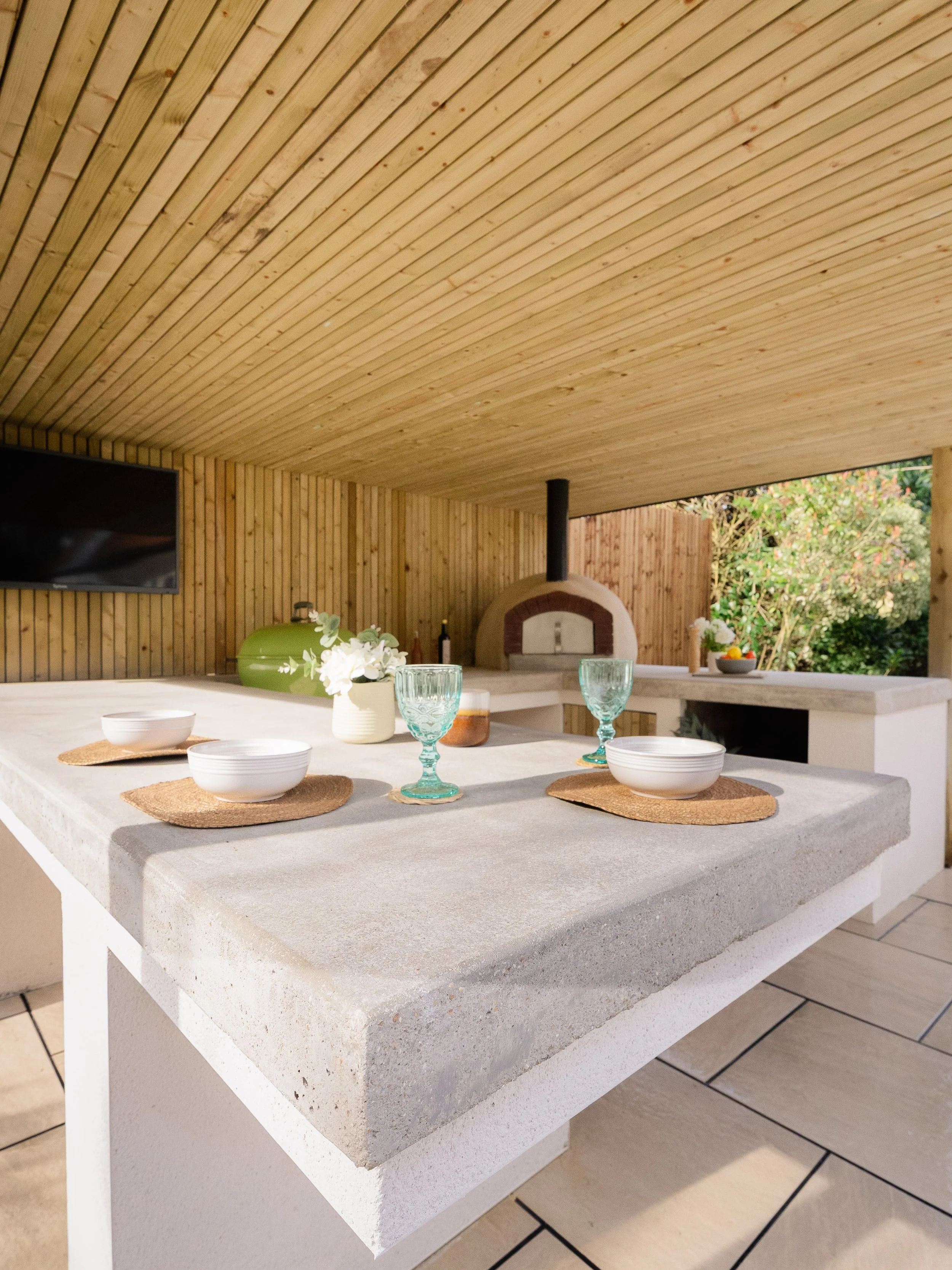 Outdoor Kitchen with a concrete worktop, set with bowls, glasses, and placemats, featuring a wood-fired pizza oven and lush greenery in the background, under a pergola with softwood cladded ceiling. Expert landscapers and garden designers in Surrey.