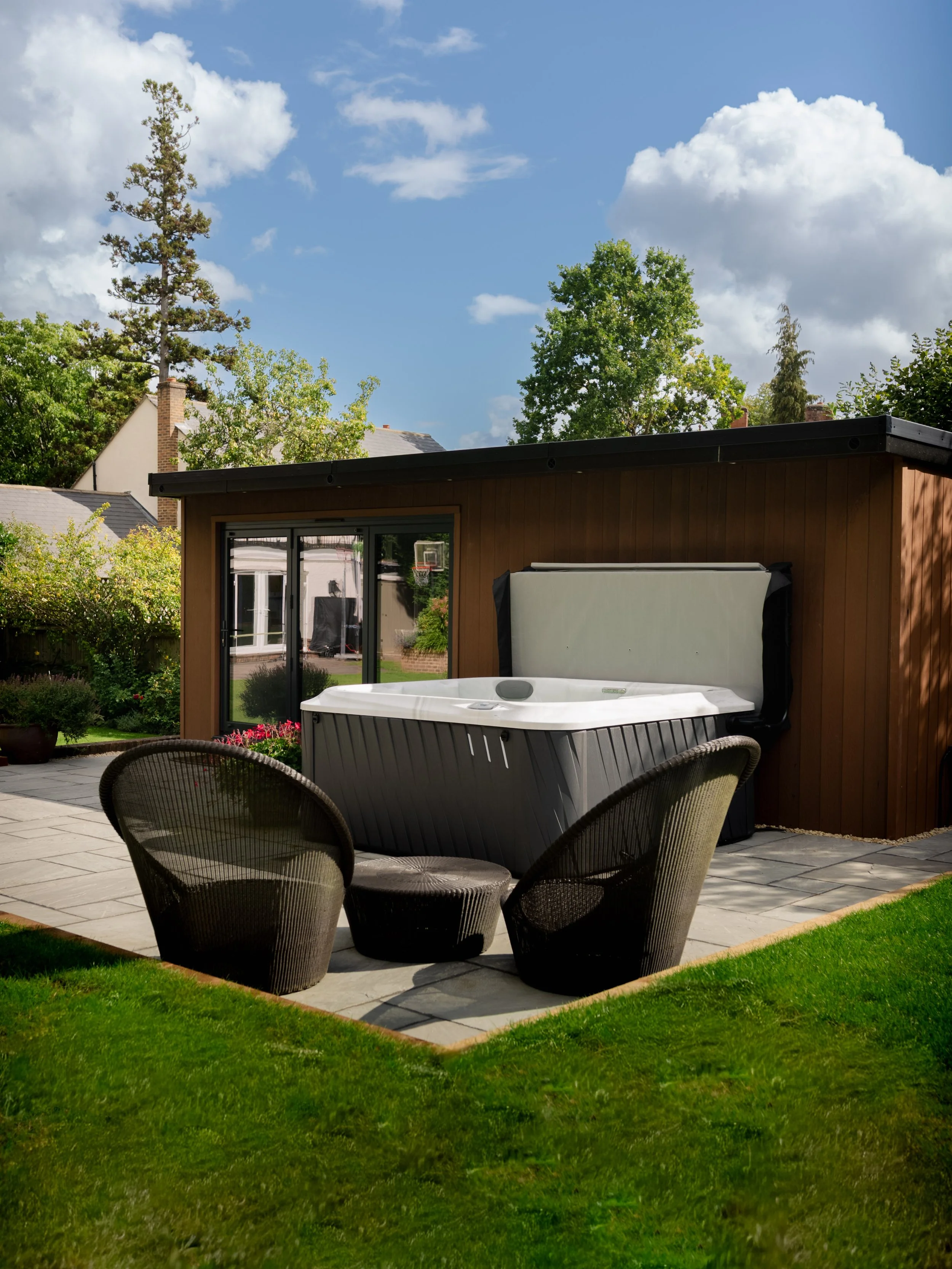 A backyard with a hot tub, patio furniture, and a wooden building under a partly cloudy sky.