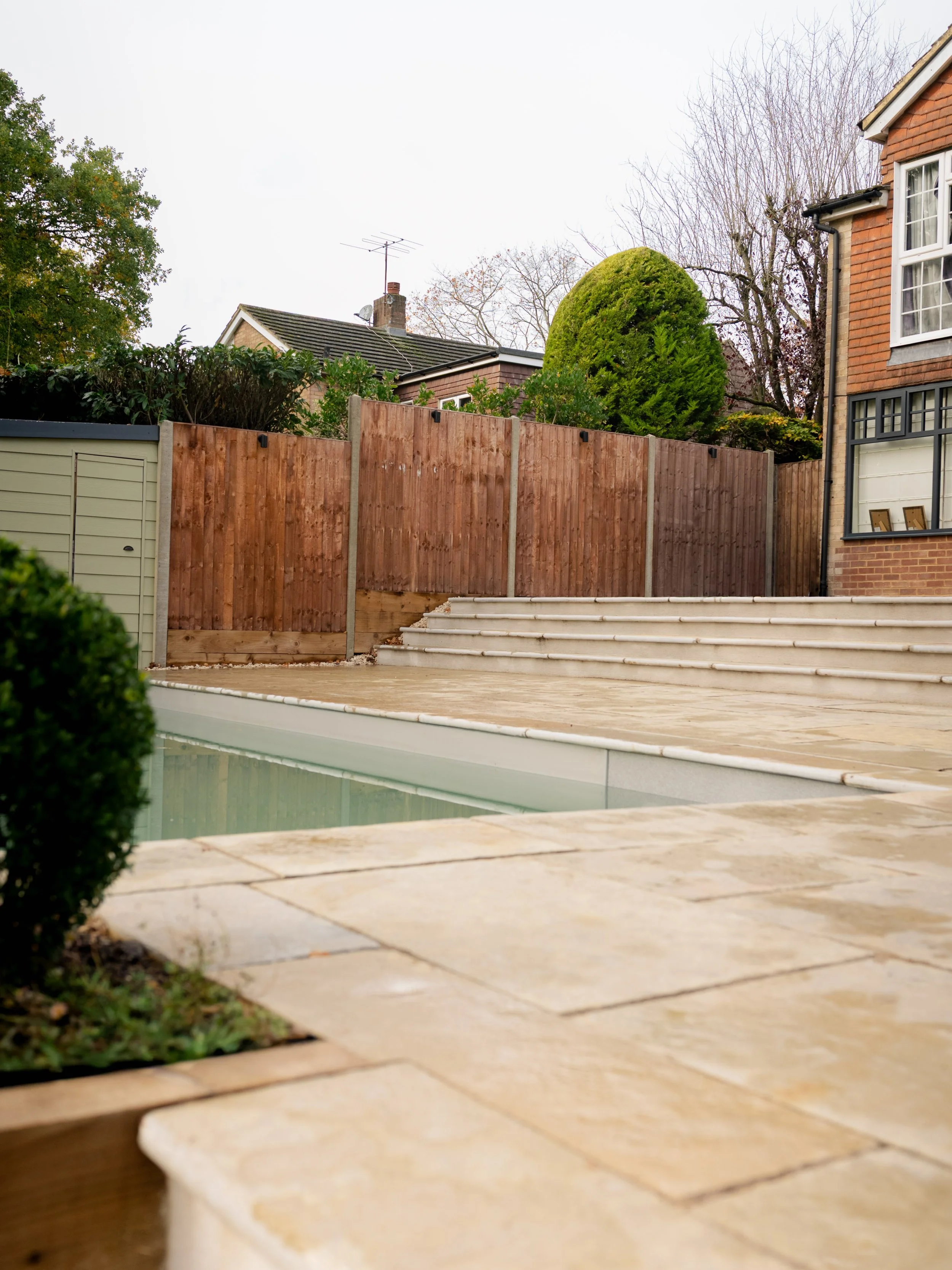 Backyard with pool, wooden deck, stairs, and a fence, cloudy sky, trees, and house wall in the background.