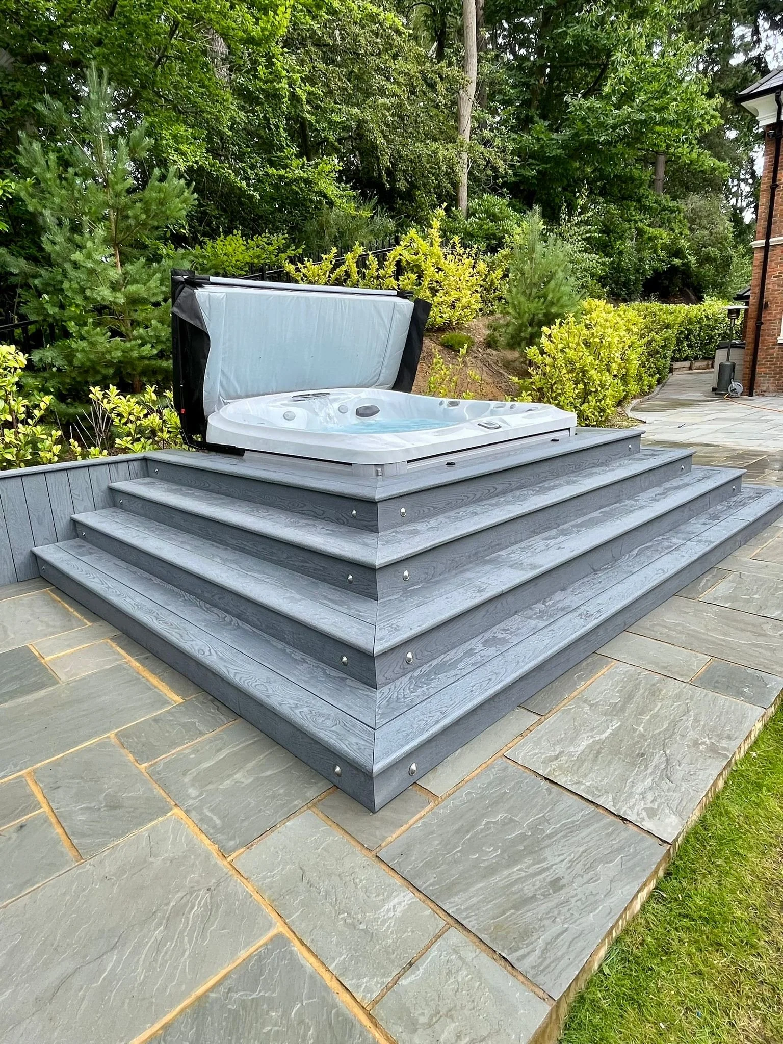 A white hot tub with a cover, situated on wooden steps outdoors, surrounded by greenery and bushes.