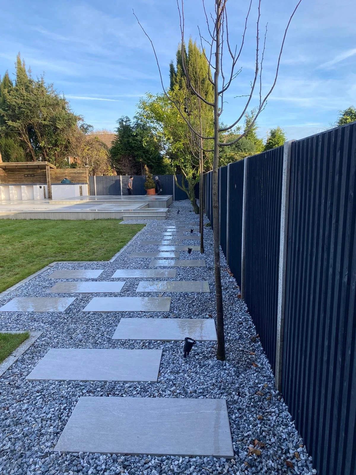 A landscaped backyard with a stone pathway, young leafless trees, a green lawn, a tall blue fence, and a deck area in the background, with two people standing and talking.