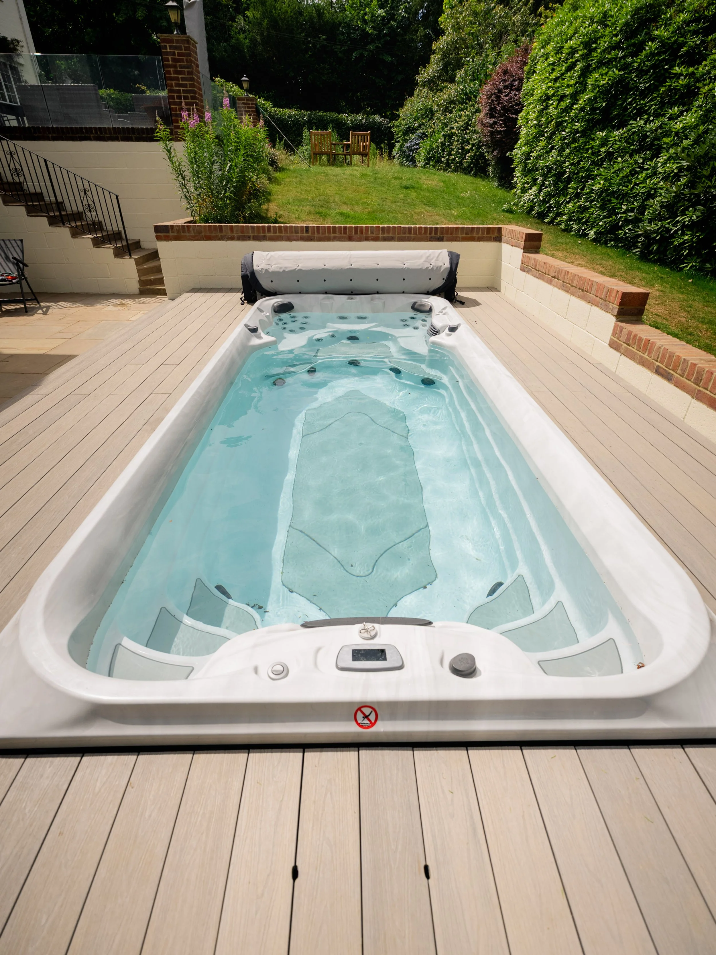 A rectangular swim spa filled with water on a wooden deck in a backyard, with an outdoor grassy area and bushes in the background. Expert landscapers and garden designers in Surrey, Hampshire, Berkshire, Camberley, London and surrounding areas.
