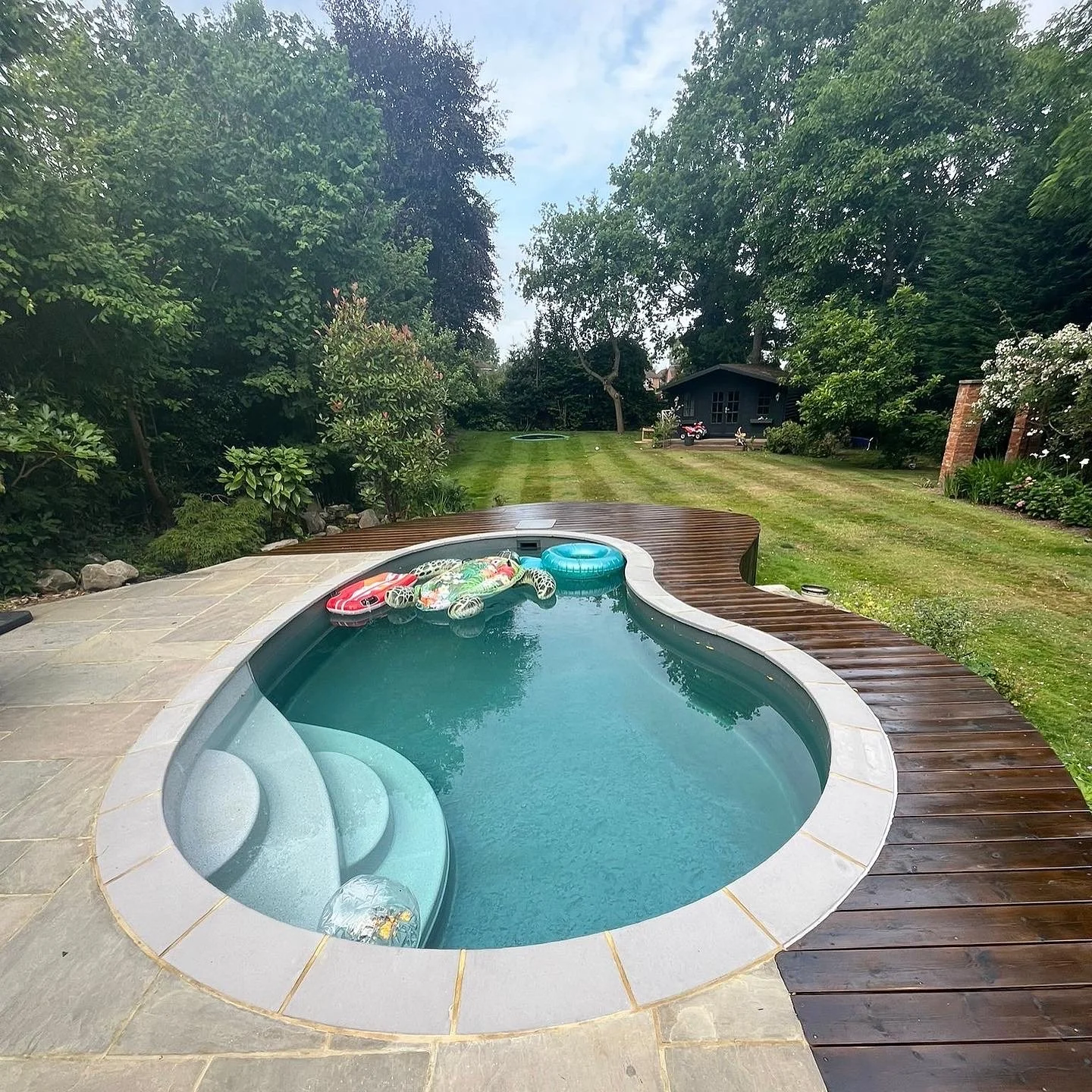 Backyard with a small kidney-shaped swimming pool, pool floaties, a towel, and a wooden deck. Green grass, trees, and a small shed in the background.