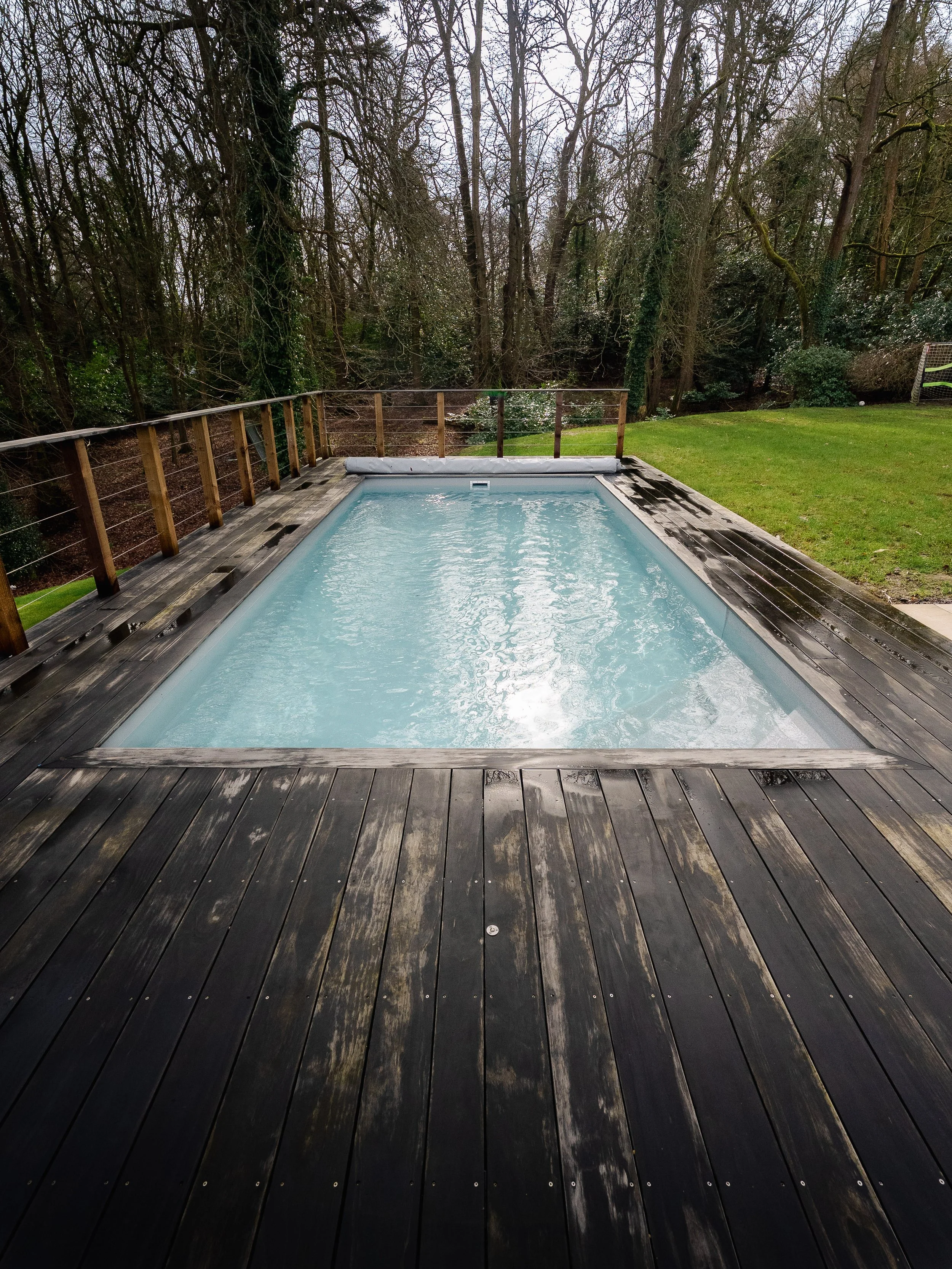 An outdoor pool with water on a wooden deck, surrounded by a grassy yard and trees.
