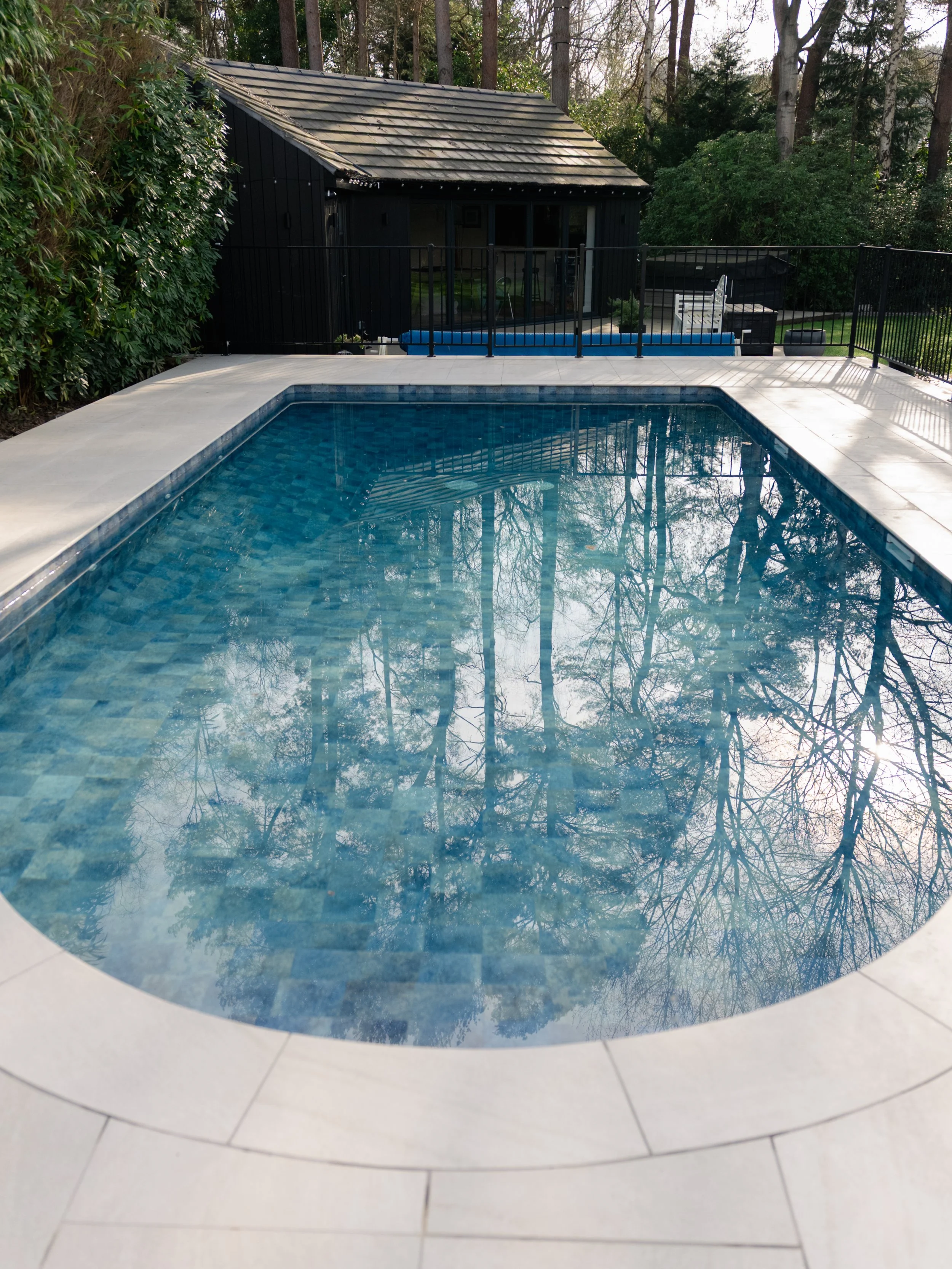 An outdoor swimming pool surrounded by a porcelain tiles and railing, with trees in the background, built by leading swimming pool builders and installers Esme Garden Design & Landscaping, a landscaping company serving surrey, hampshire, berkshire, a