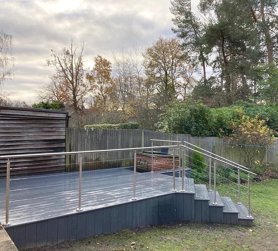Backyard with a small elevated deck, staircase, metal railing, and a wooden fence, with trees in the background under a cloudy sky.