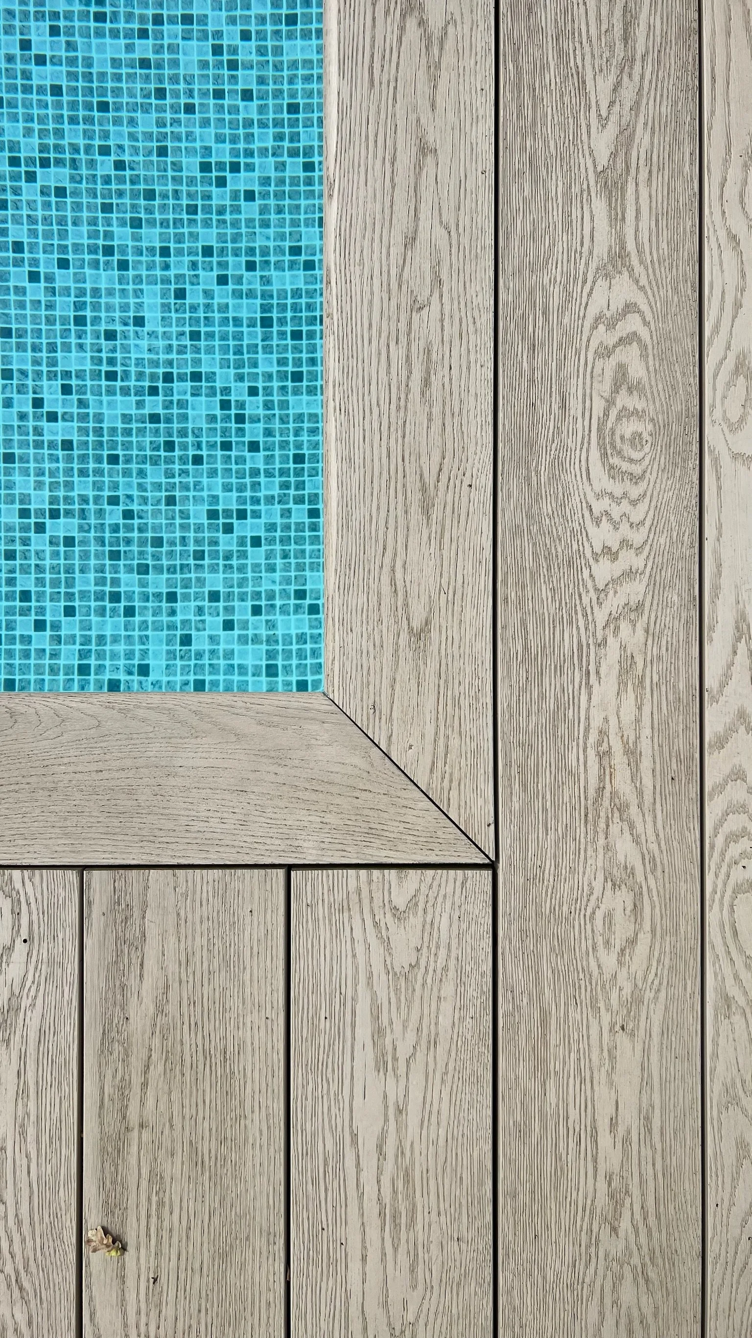 Close-up of a corner of a wooden deck with light grey planks and a section of blue mosaic tiles.