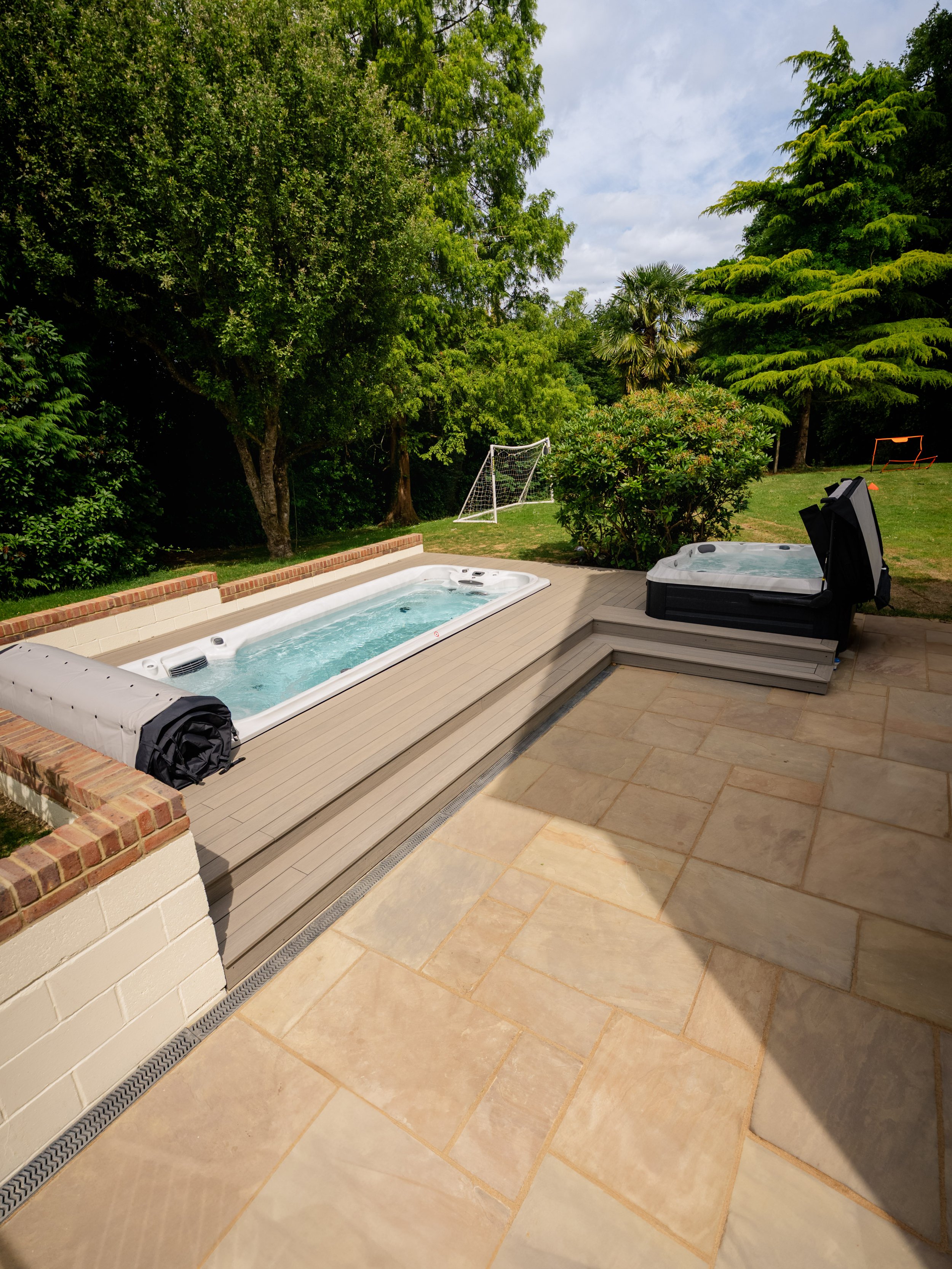 A garden with a hot tub on a patio, surrounded by lush green trees and a grassy area with a small soccer goal. Expert landscapers and garden designers in Surrey, Hampshire, Berkshire, Camberley, London and surrounding areas.