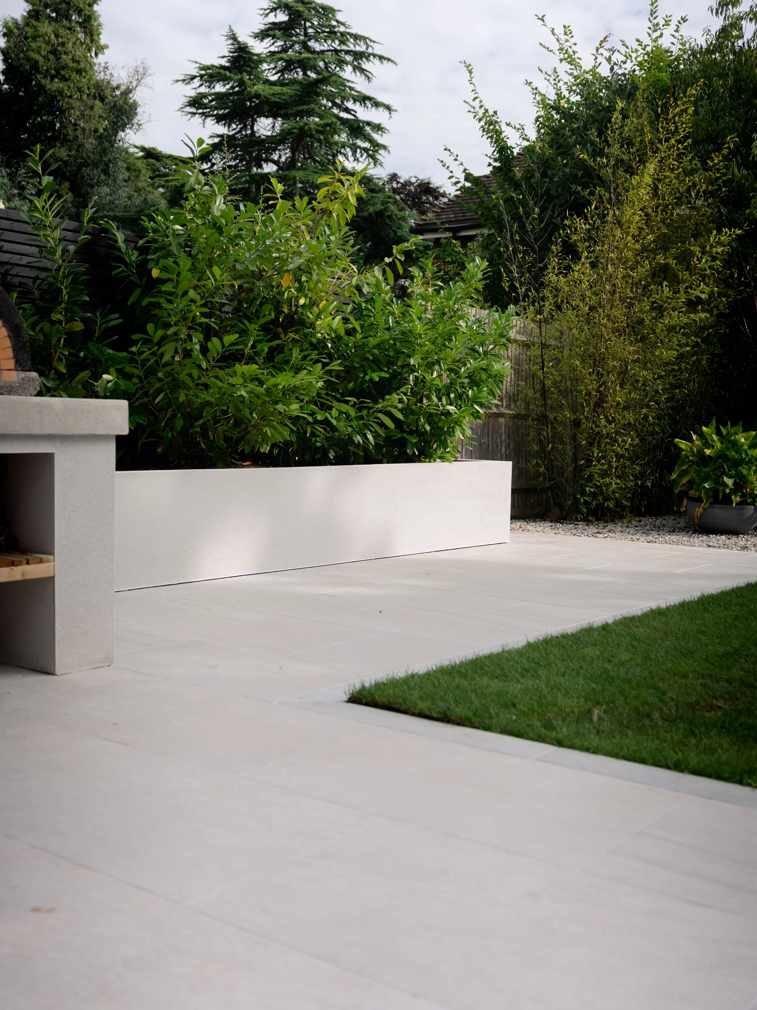 Modern garden with white tiled patio, green bushes, trees, and a small patch of grass. Expert landscapers and garden designers in Surrey, Hampshire, Berkshire, Camberley, London and surrounding areas.