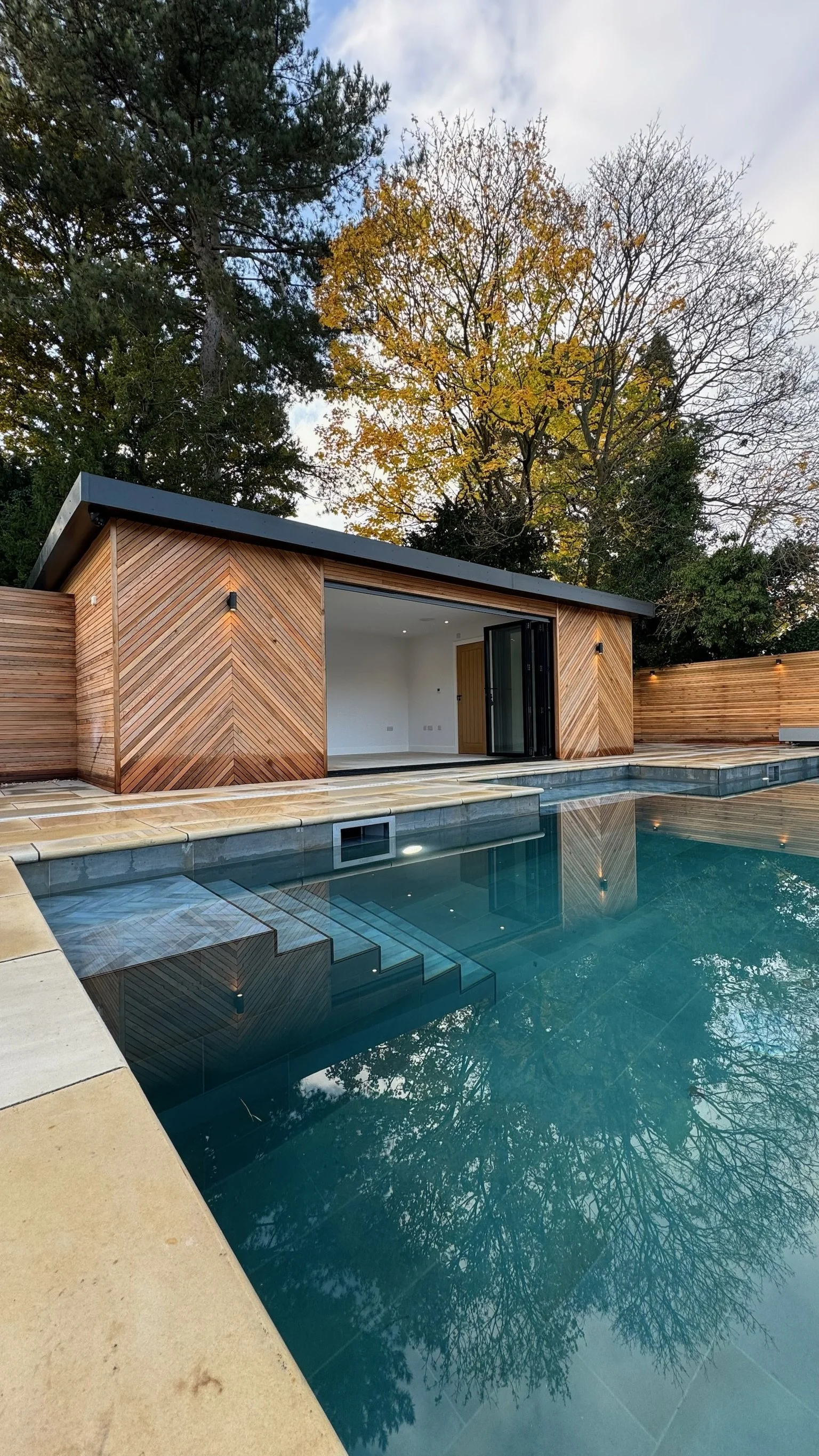 Modern backyard pool with a small wooden pool house and a privacy wooden fence, surrounded by trees with autumn leaves.