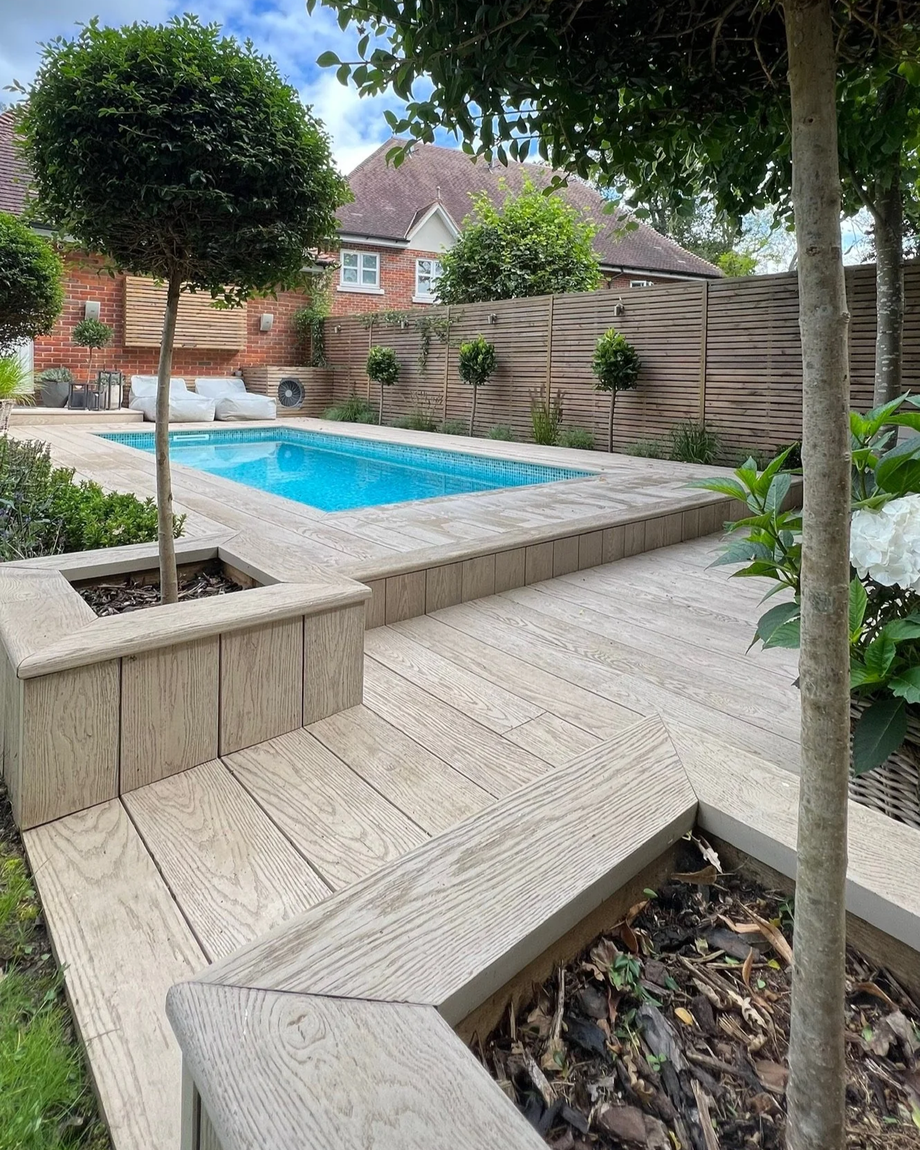 A garden with a swimming pool, surrounded by a wooden deck, some trees, and a fence with green plants. Expert landscapers and garden designers in Surrey, Hampshire, Berkshire, Camberley, London and surrounding areas.