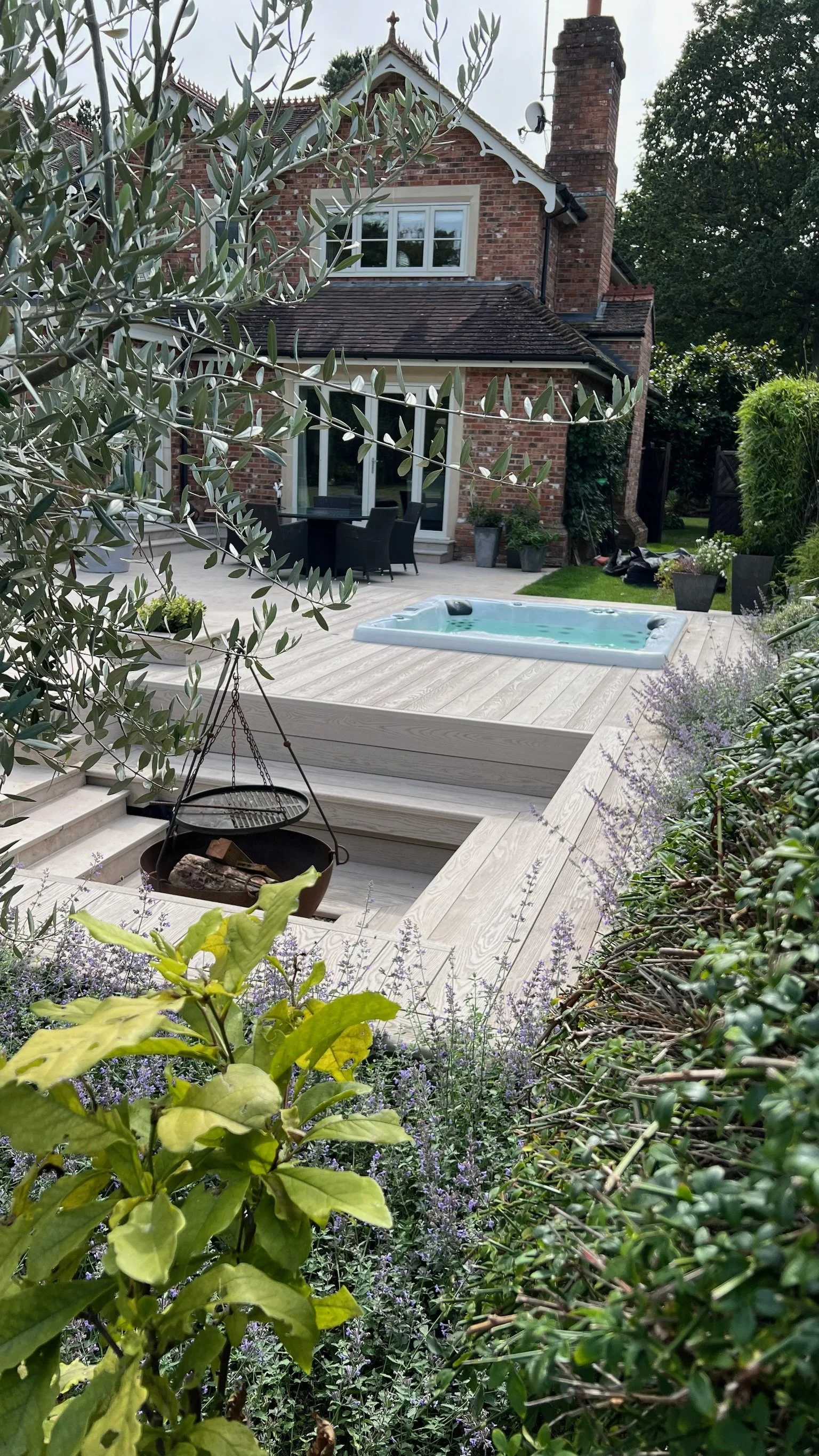 A garden with a millboard deck, hot tub, and firepit area, surrounded by plants. Esme is a garden design & landscaping company serving surrey, hampshire, berkshire, camberley, london & surrounding areas. We build bespoke outdoor living spaces. 