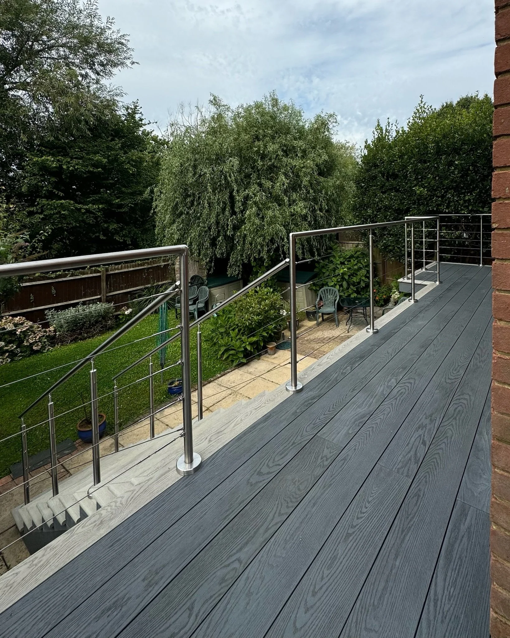 A modern balcony with millboard decking and metal railings overlooking a garden with green trees, bushes, and outdoor furniture. Esme are a widely trusted garden design and landscaping company serving surrey, hampshire, berkshire, ascot, london.