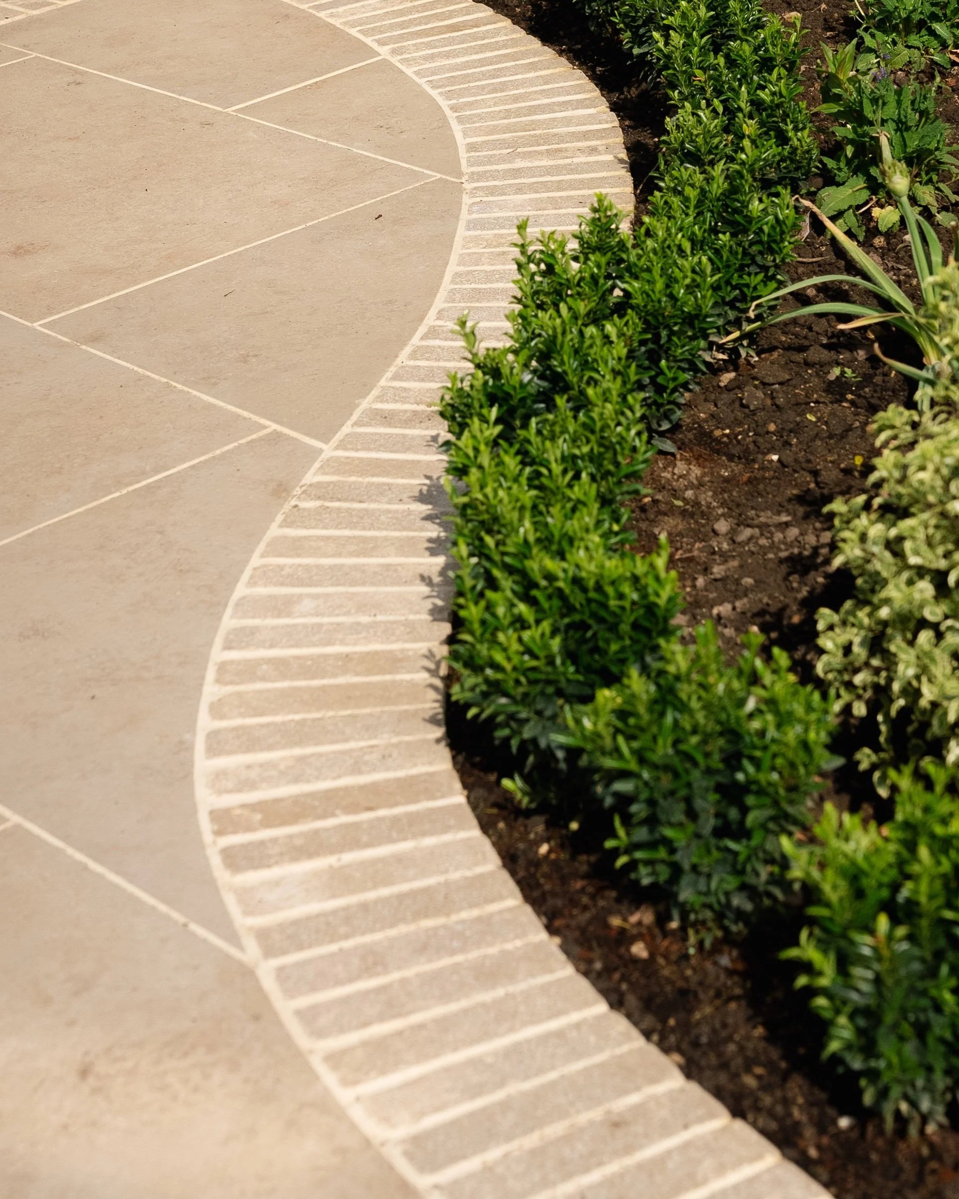 Curved brick border beside a concrete sidewalk with green plants and soil on the right side. Expert landscapers and garden designers in Surrey, Hampshire, Berkshire, Camberley, London and surrounding areas.