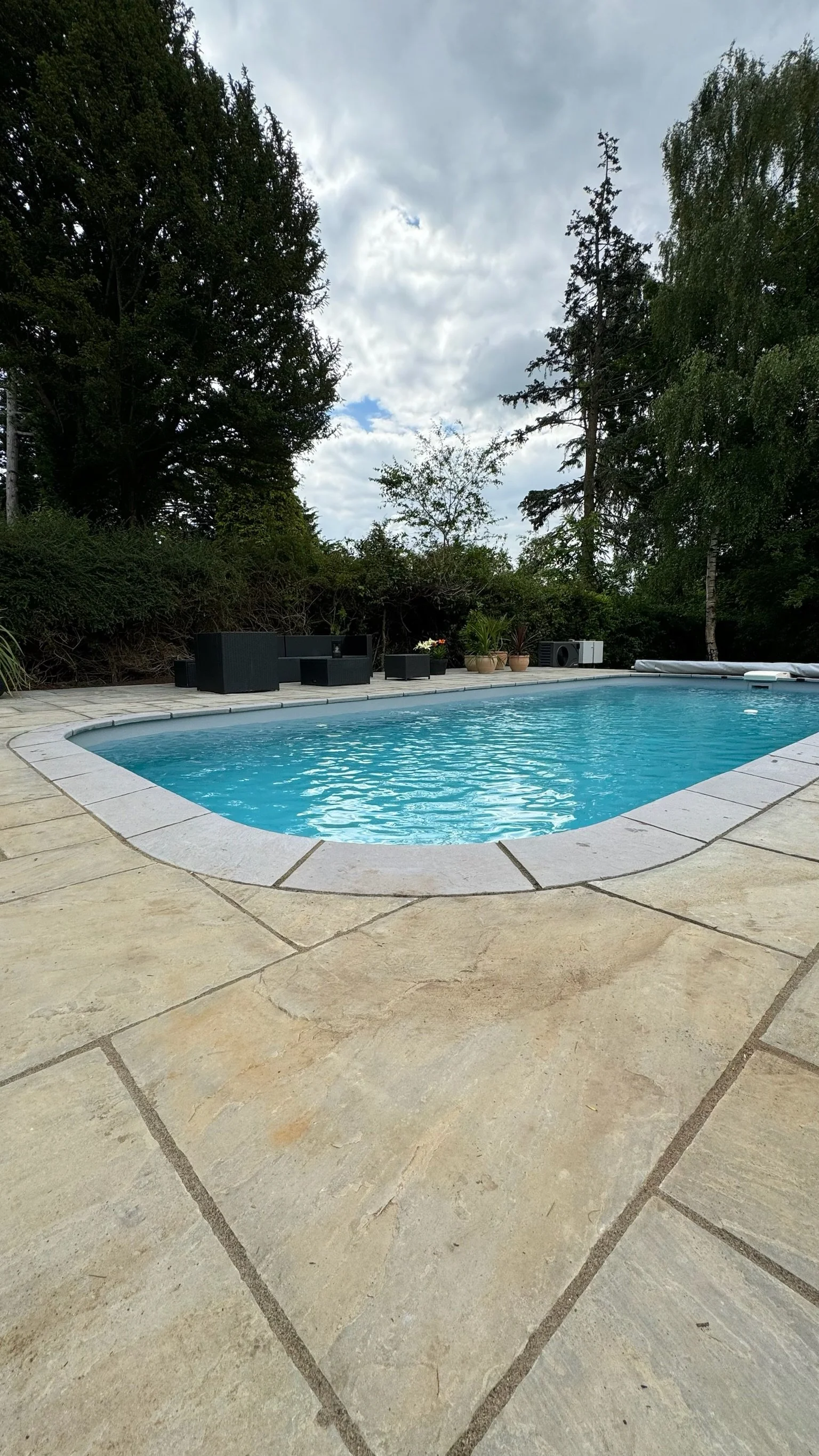 A backyard pool surrounded by stone tiles with a seating area and trees in the background under a cloudy sky.