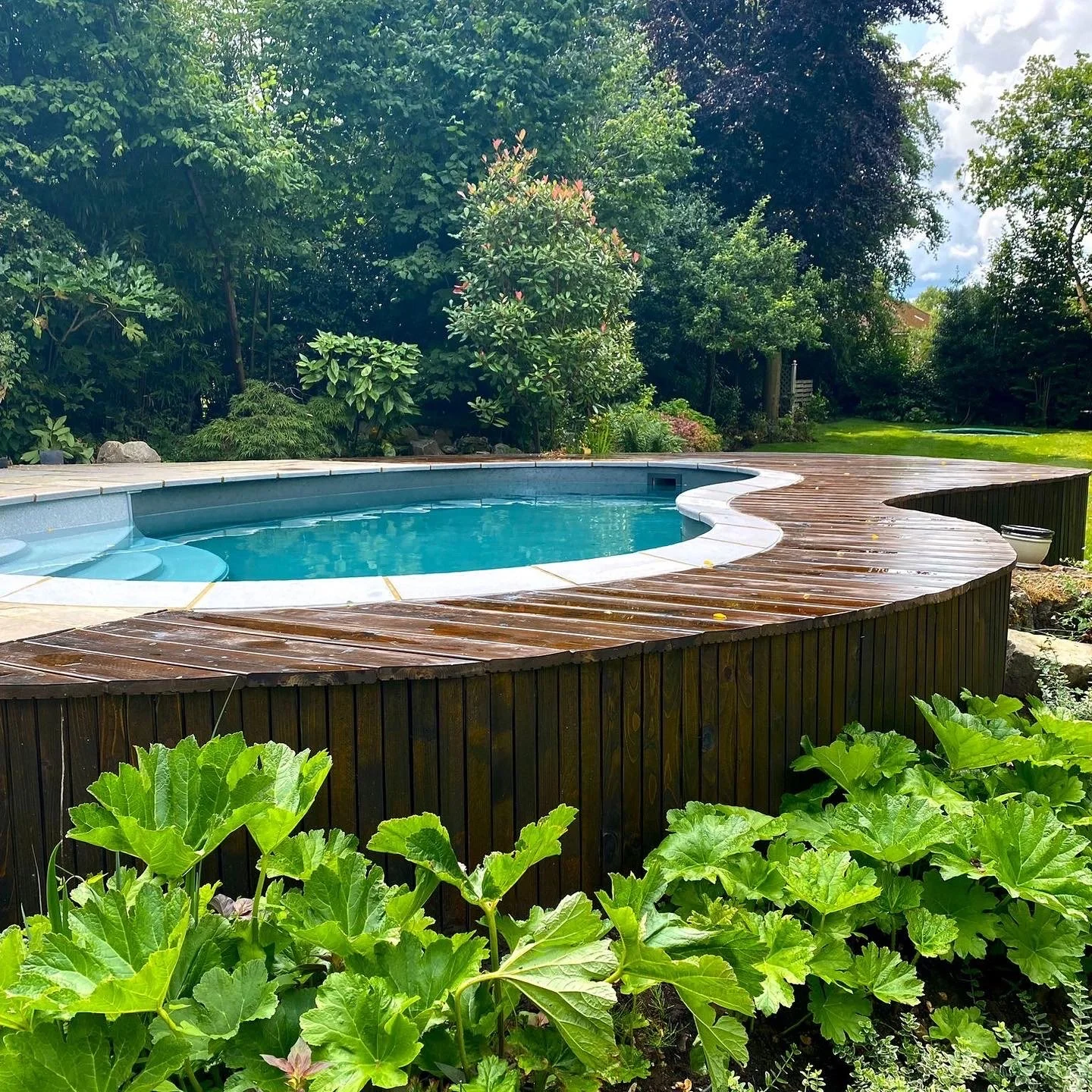 A backyard swimming pool surrounded by a wooden deck and lush green trees and plants.