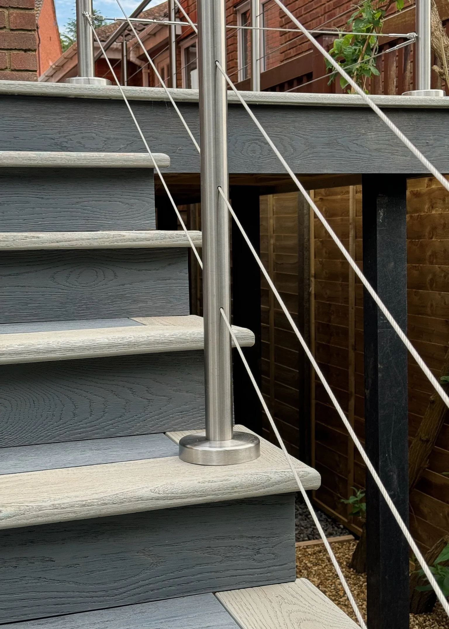 Close-up of outdoor wooden stairs with metal railing and cable wires, with a wooden fence and some plants in the background. Esme are a widely trusted garden design and landscaping company serving surrey, hampshire, berkshire, ascot, london.
