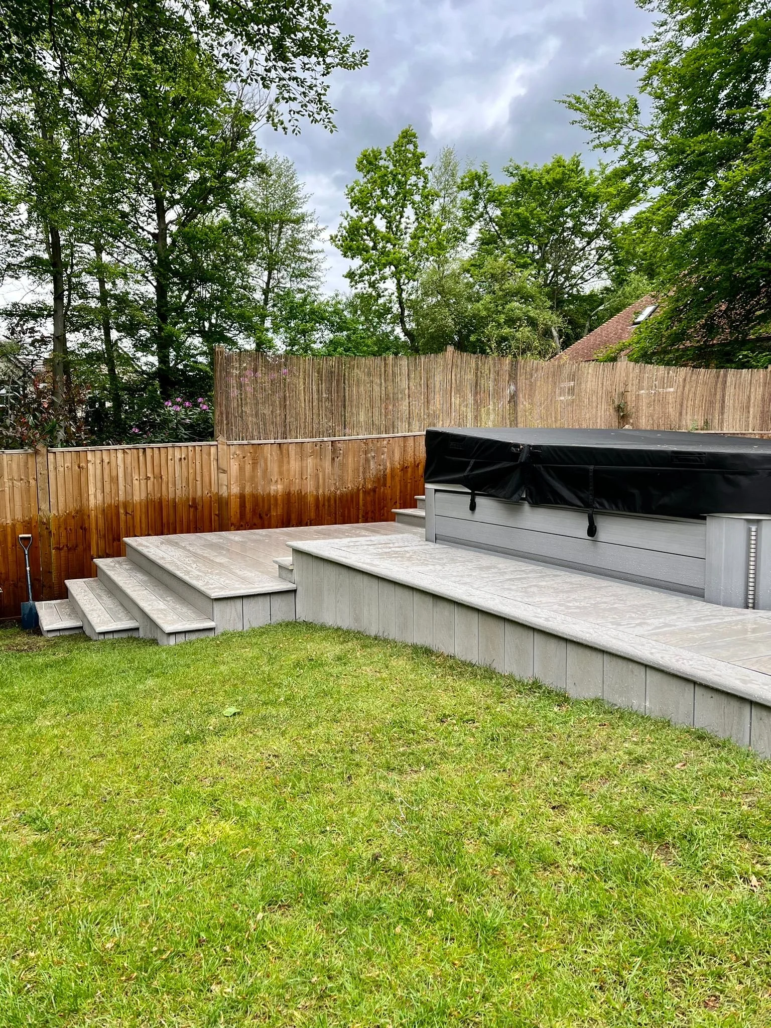 A backyard with a grass lawn, a wooden deck with stairs, a hot tub covered with a black cover, and a bamboo privacy fence, surrounded by lush green trees under a cloudy sky.
