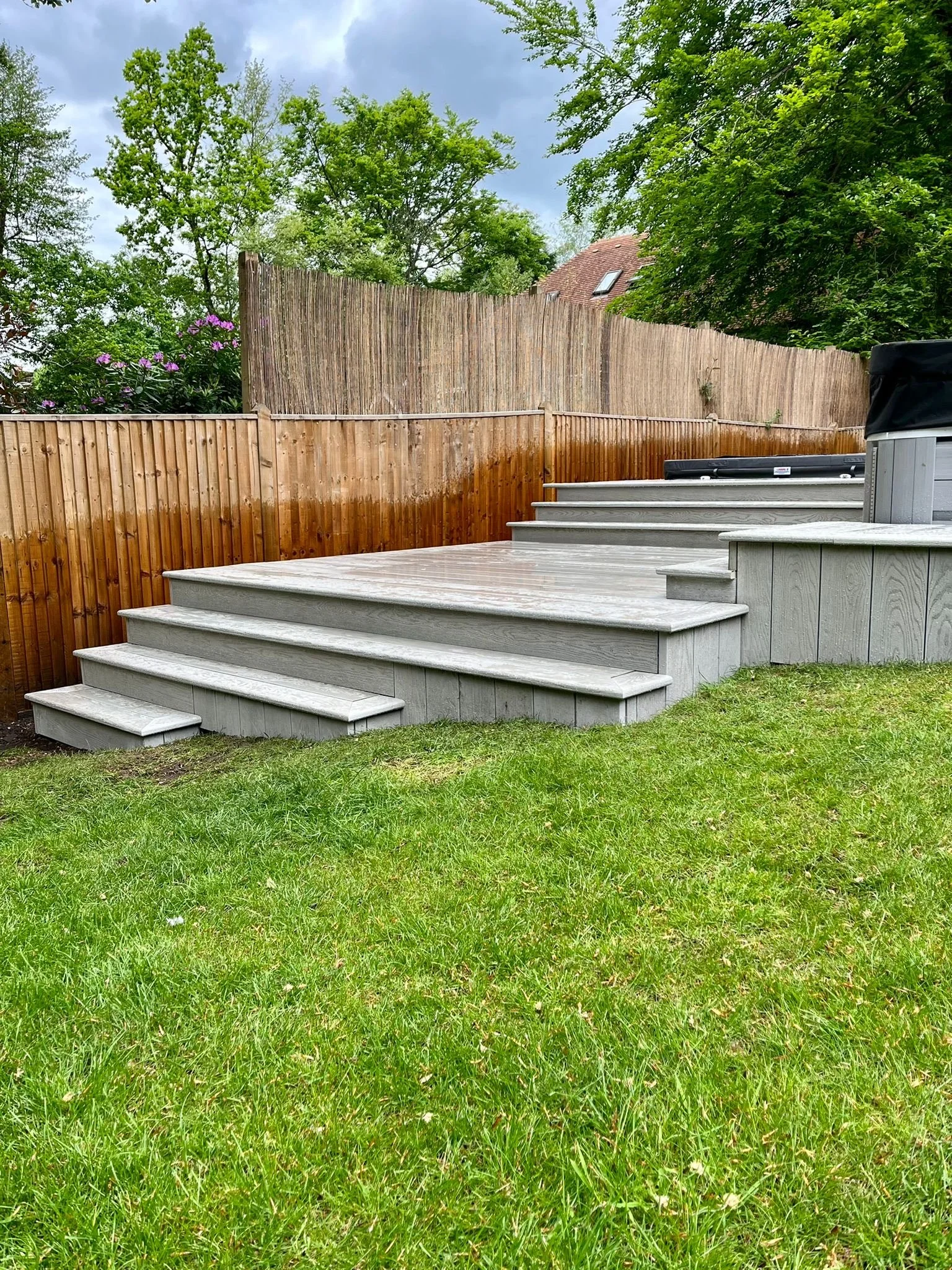 A backyard with a green lawn and a raised wooden deck with stairs, enclosed by a wooden fence and tall trees.