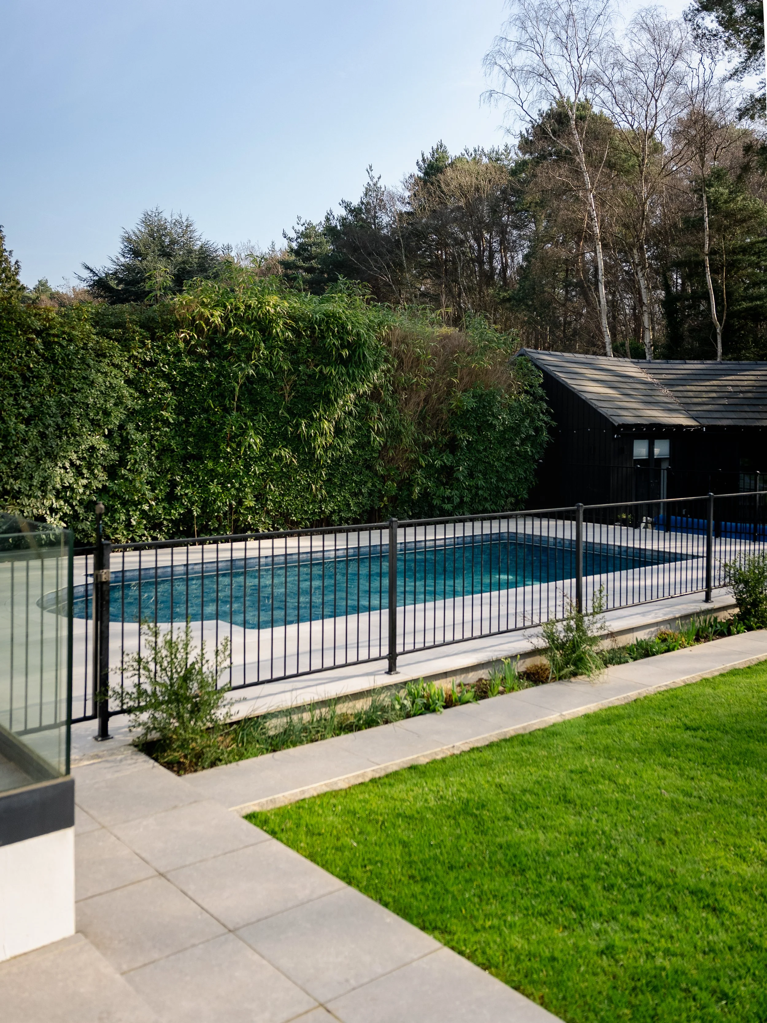 An outdoor swimming pool surrounded by a porcelain tiles and railing, with trees in the background, built by leading swimming pool builders and installers Esme Garden Design & Landscaping, a landscaping company serving surrey, hampshire, berkshire, a