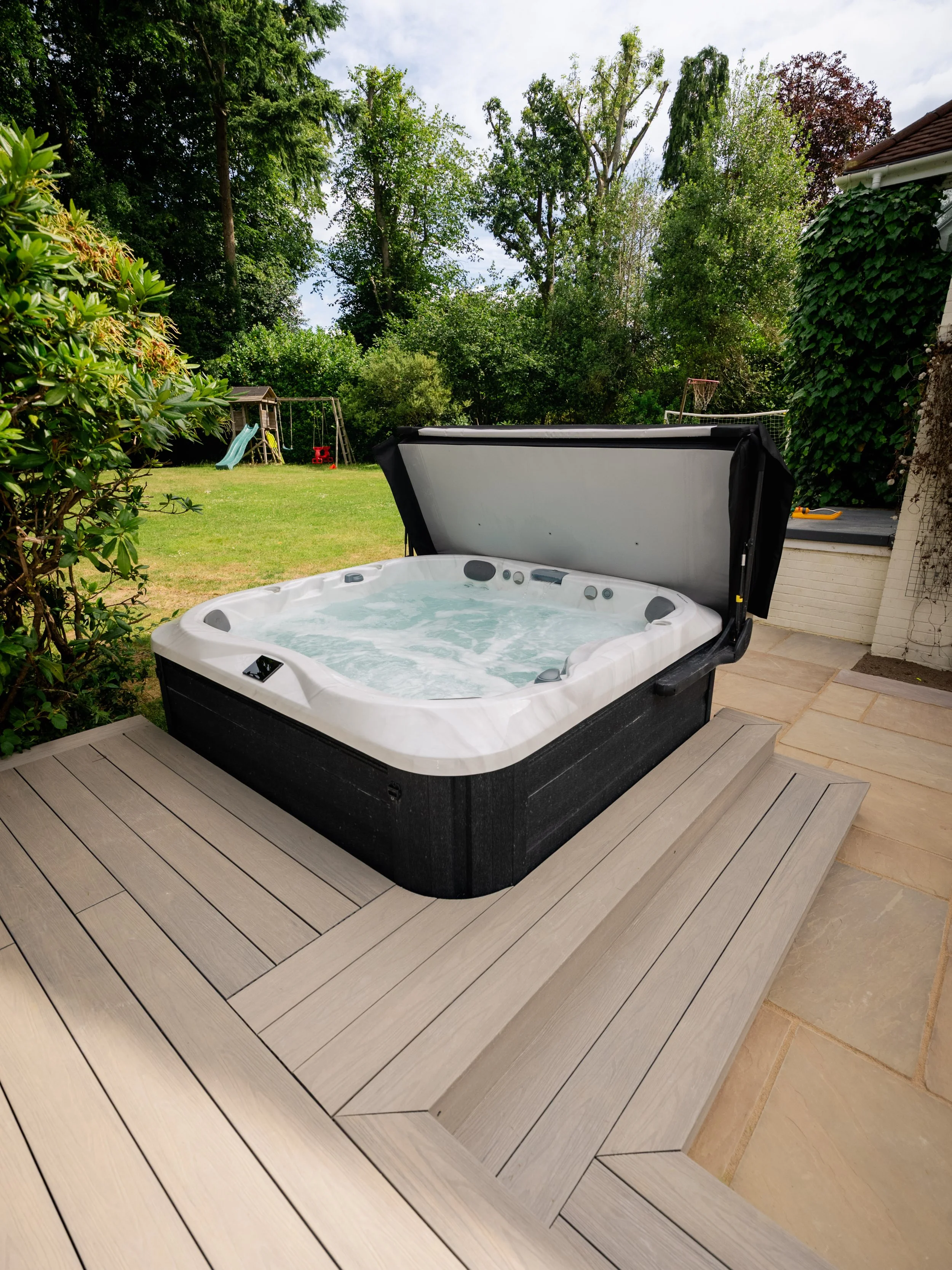 Outdoor hot tub with opened cover on a wooden deck, surrounded by green trees and a backyard with a slide and playground in the background. Expert landscapers and garden designers in Surrey, Hampshire, Berkshire, Camberley, London and surrounding are
