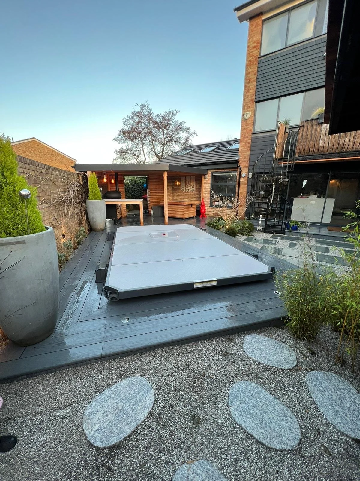 A backyard patio with a covered seating area, potted plants, an outdoor dining set, a spiral staircase, and a hot tub cover on the wooden deck, with a house and trees in the background.