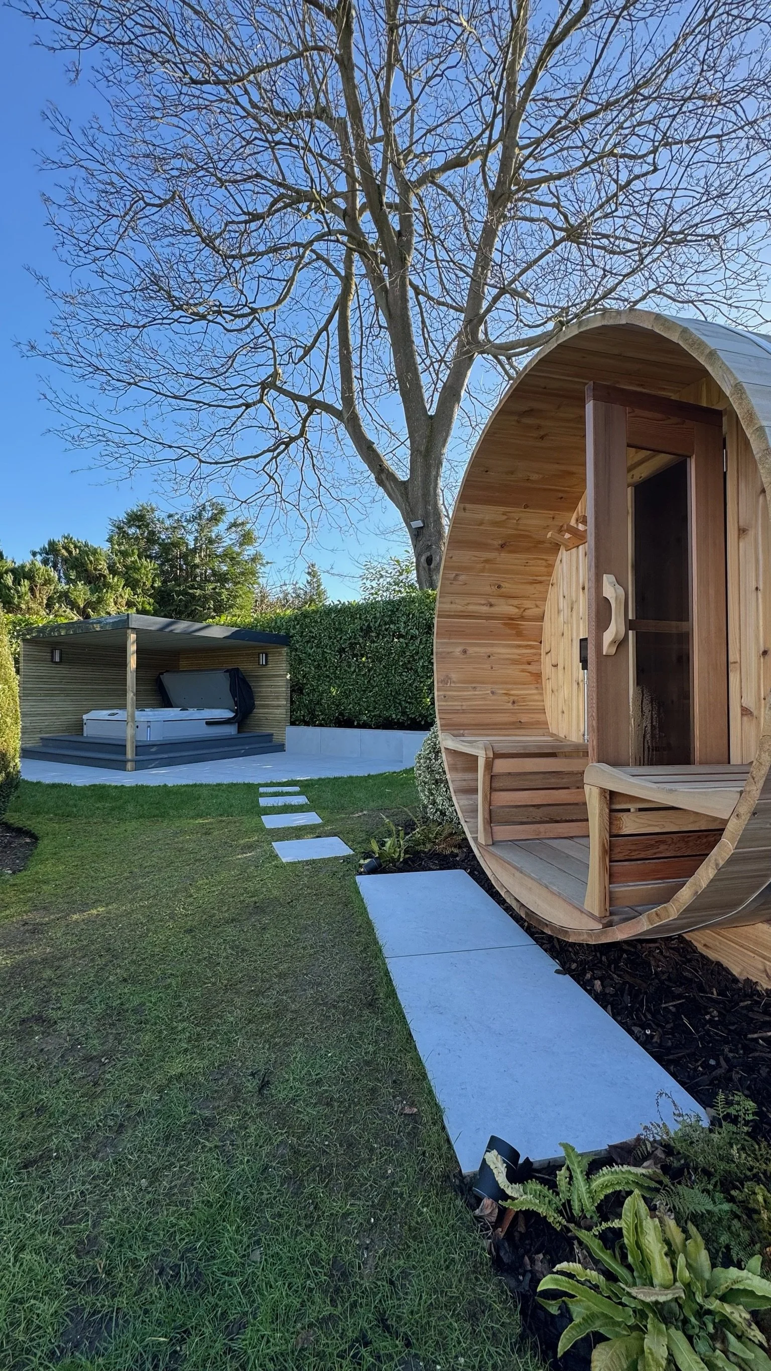 Garden with a outdoor sauna pod made of wood, a hot tub with a cover, a stepping stone pathway. Esme is a garden design & landscaping company serving surrey, hampshire, berkshire, camberley, london & surrounding areas. We build bespoke gardens. 