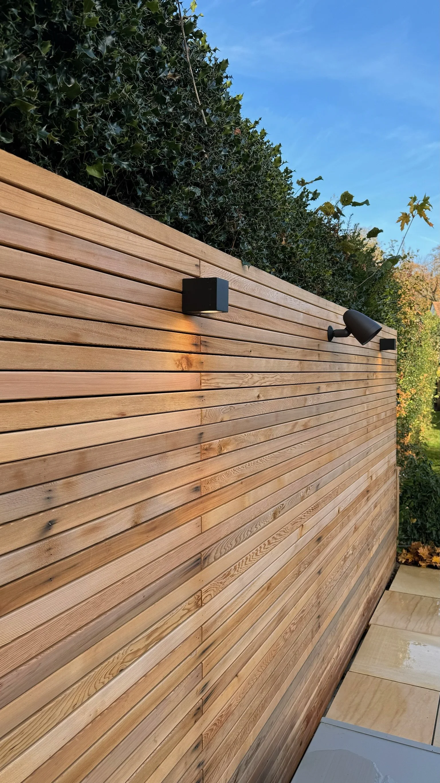 Exterior view of a wooden privacy fence with black outdoor light fixtures against a blue sky with some trees in the background.