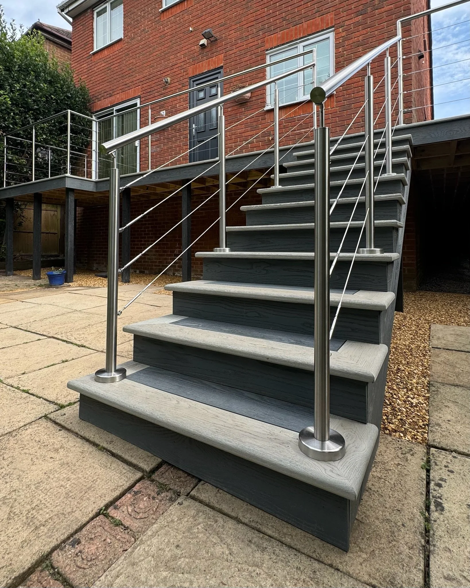 The image shows one of Esme's decking and balustrading projects, an outdoor staircase leading to a front door of a brick house. The staircase has millboard steps with a modern metal railing.