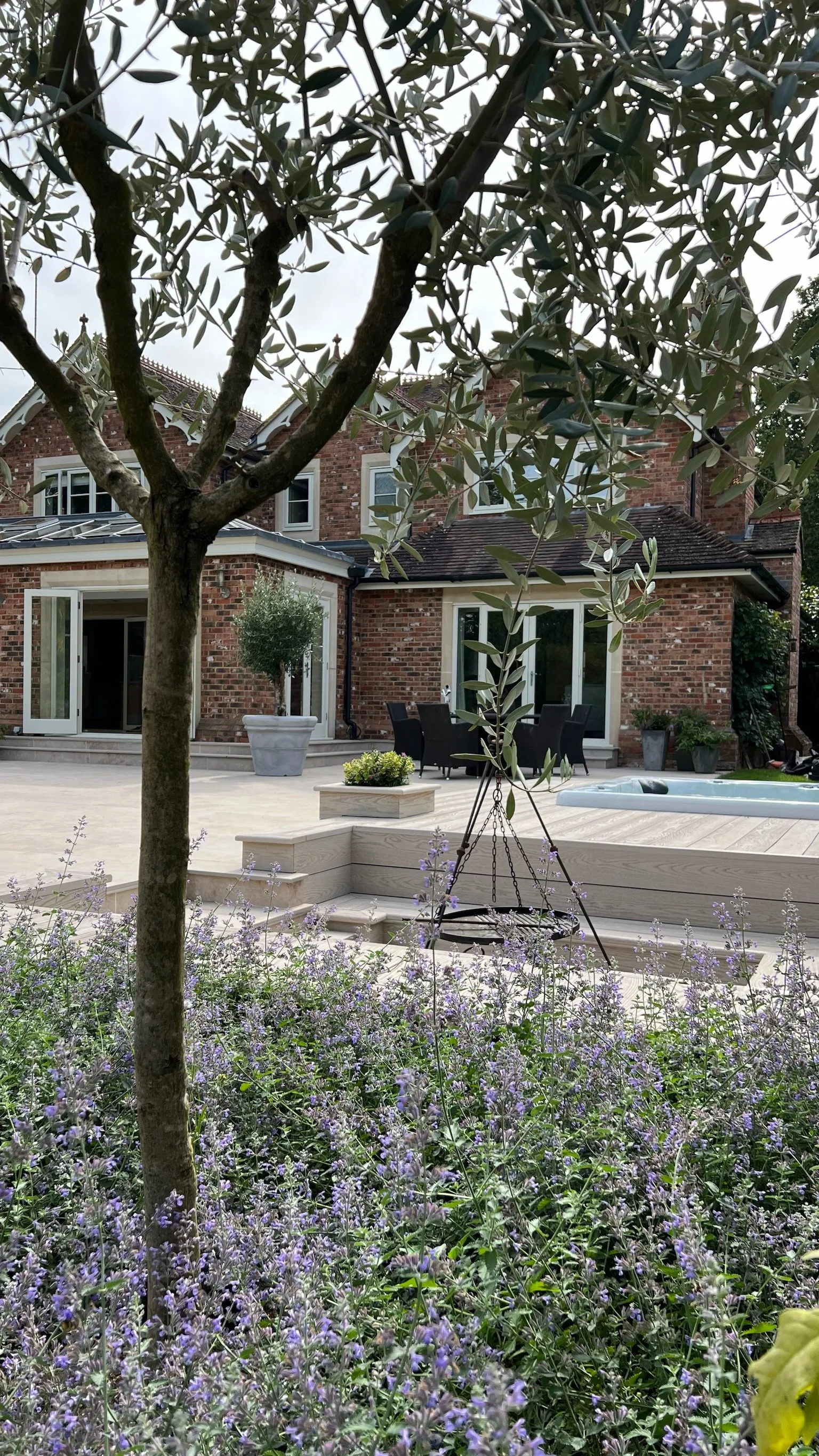 Backyard patio with flowering plants, trees, a hot tub, outdoor dining set, potted plants, and a brick house with multiple windows and doors.