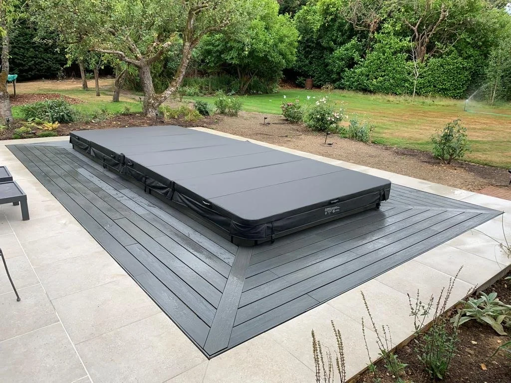 A hot tub with cover on a wooden deck in a backyard garden with trees, bushes, and a lawn.