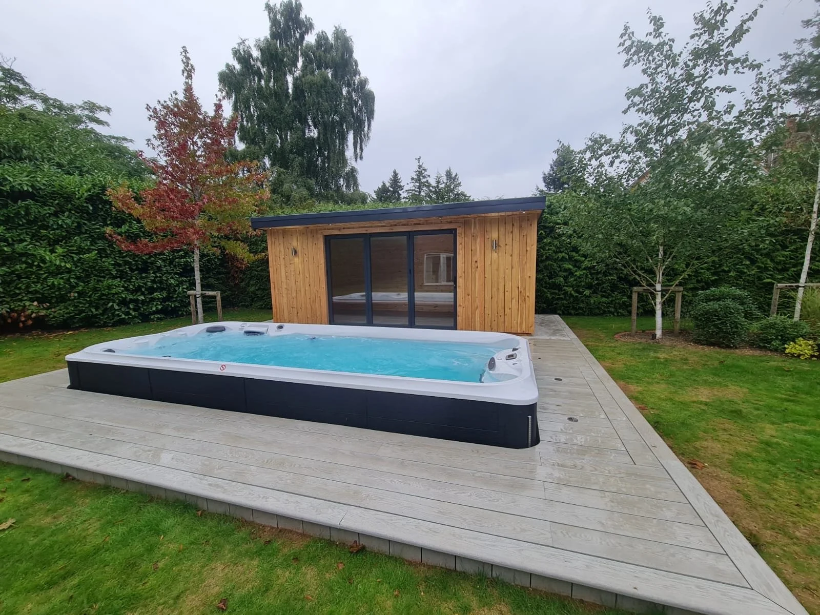 An outdoor hot tub with a black exterior is on a wooden deck surrounded by a lush green garden with trees and bushes, and a small wooden shed with sliding glass doors is in the background during overcast weather.