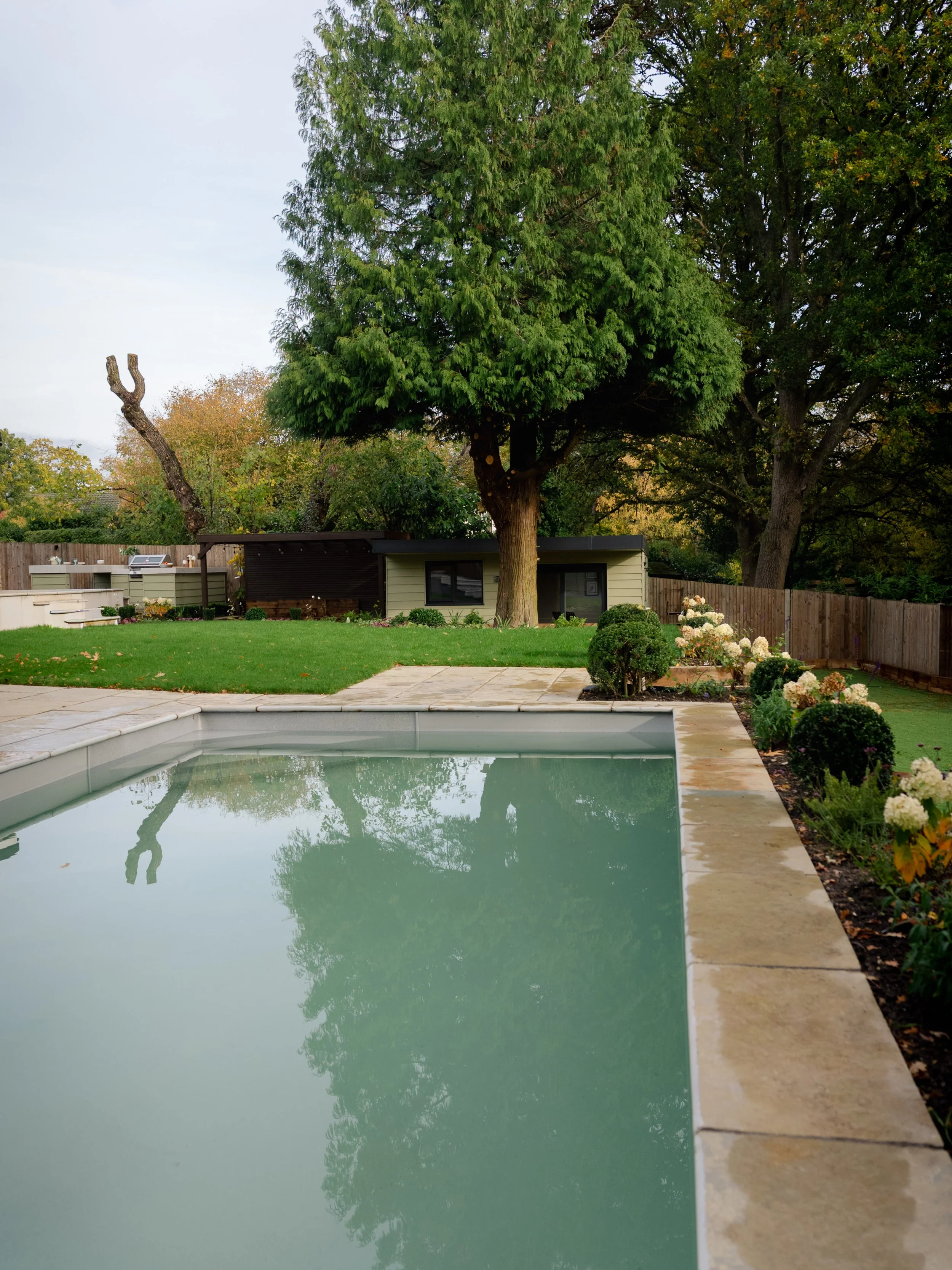 A backyard scene featuring a swimming pool, a lush green lawn, and various trees and shrubs, with a small building in the background.