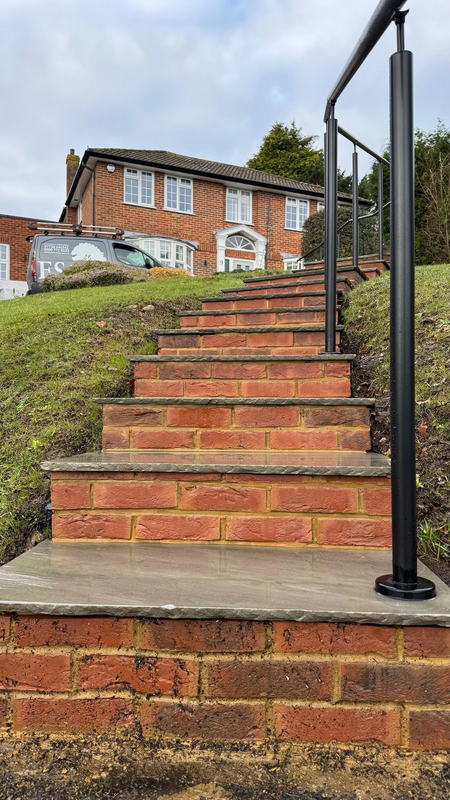 Exterior brick staircase leading up to a house with a black handrail on the right side and a lawn on either side. Esme are a widely trusted garden design and landscaping company serving surrey, hampshire, berkshire, ascot, london and surroundings
