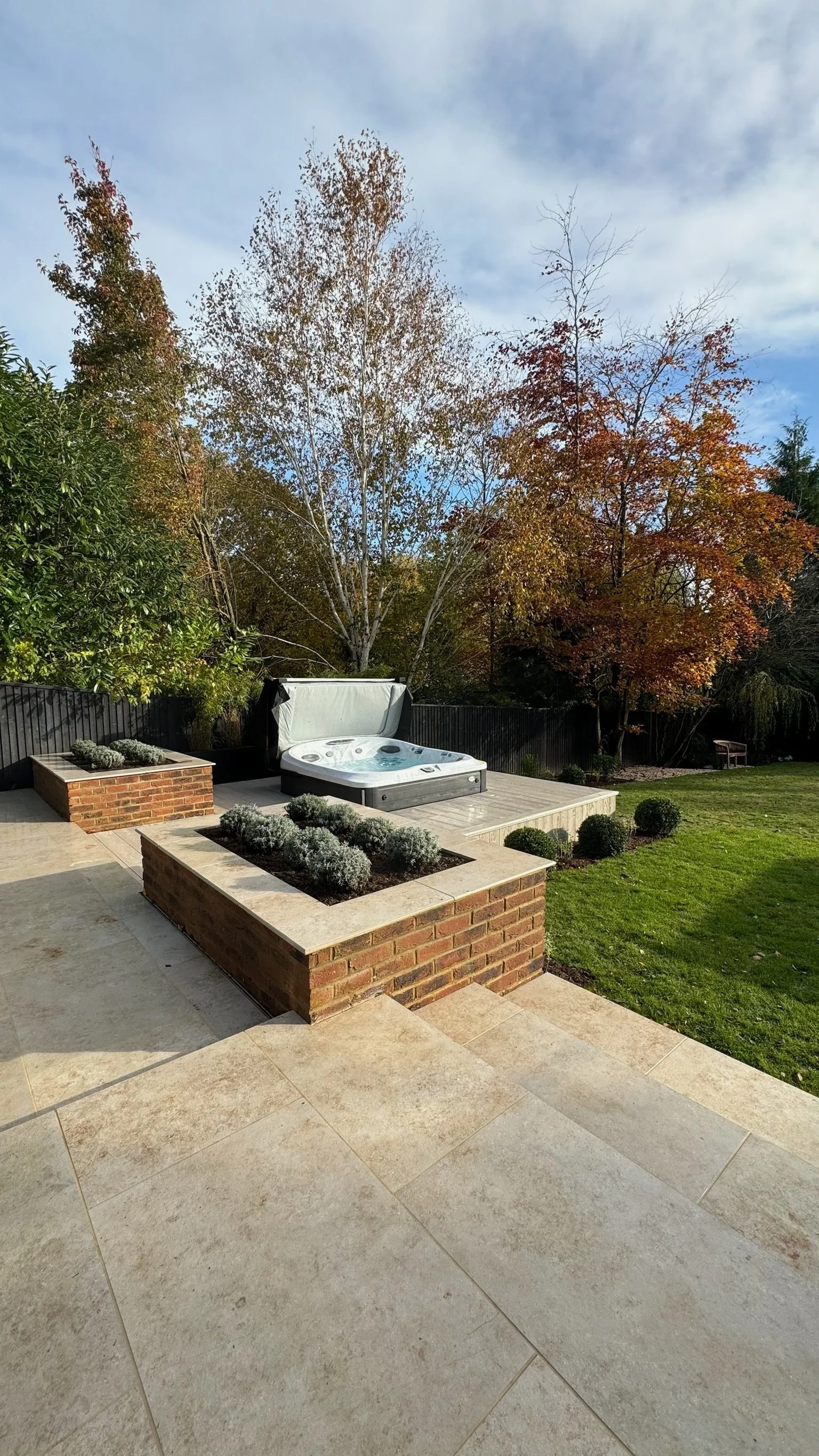Backyard with patio, raised brick flower beds with bushes, a hot tub, and trees with fall foliage under a cloudy sky.