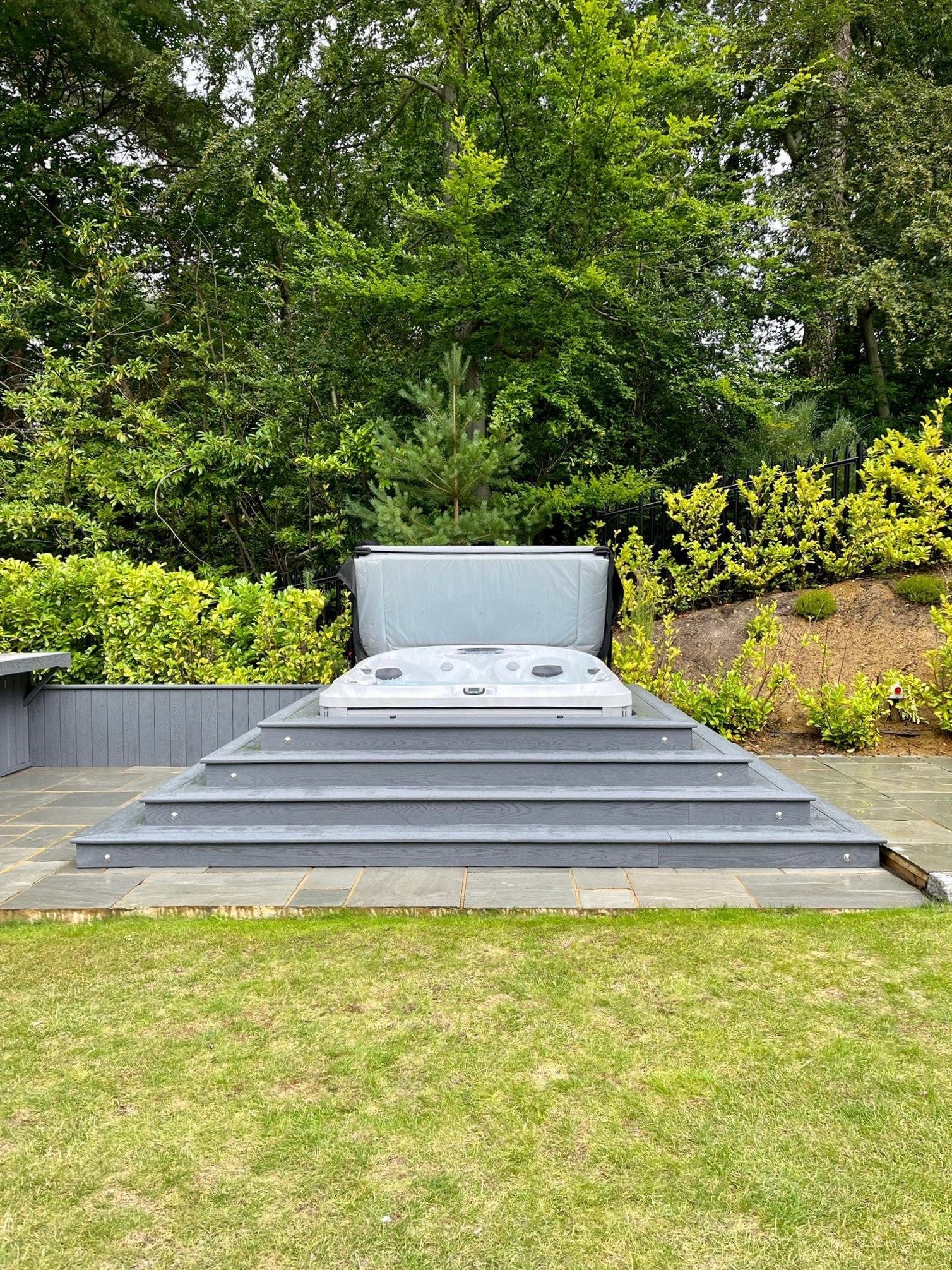 Hot tub with a cover on a deck, surrounded by greenery and trees.