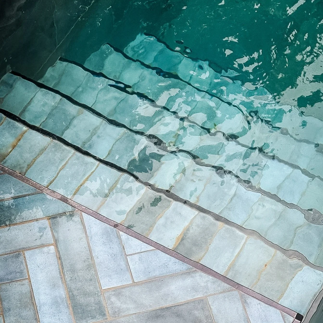 A set of tiled stairs leading into a swimming pool, with the pool water visible and rippled. Expert landscapers and garden designers in Surrey, Hampshire, Berkshire, Camberley, London and surrounding areas.