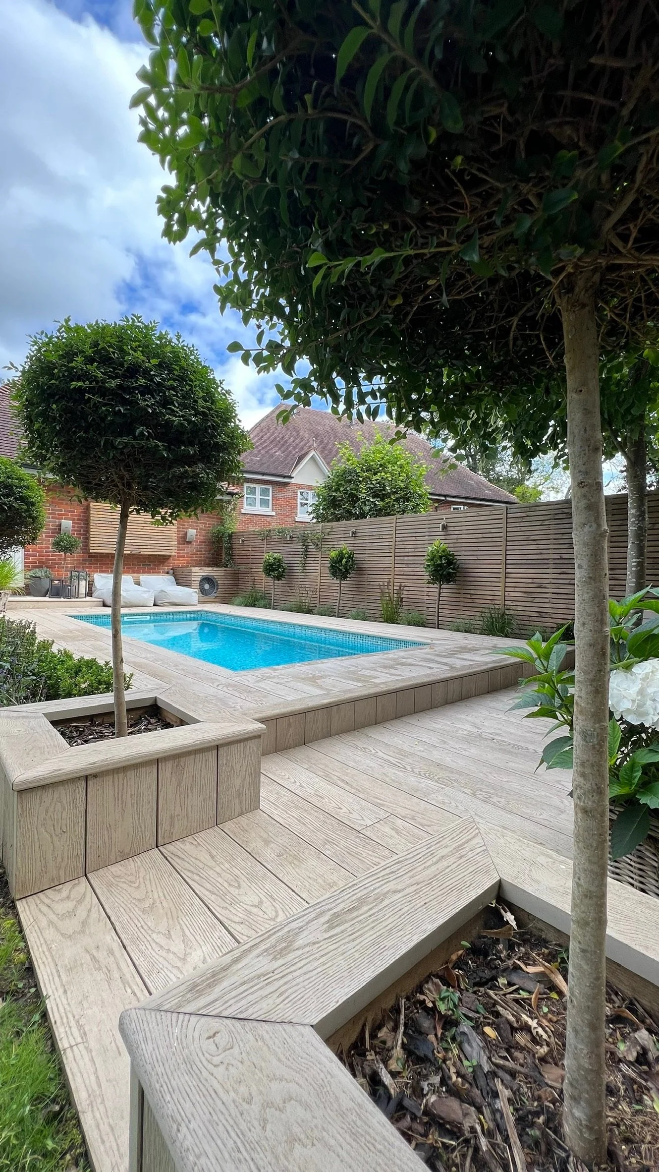 A backyard with a rectangular swimming pool, surrounded by wooden decking, potted trees, and some greenery, with a brick house and a wooden fence in the background.