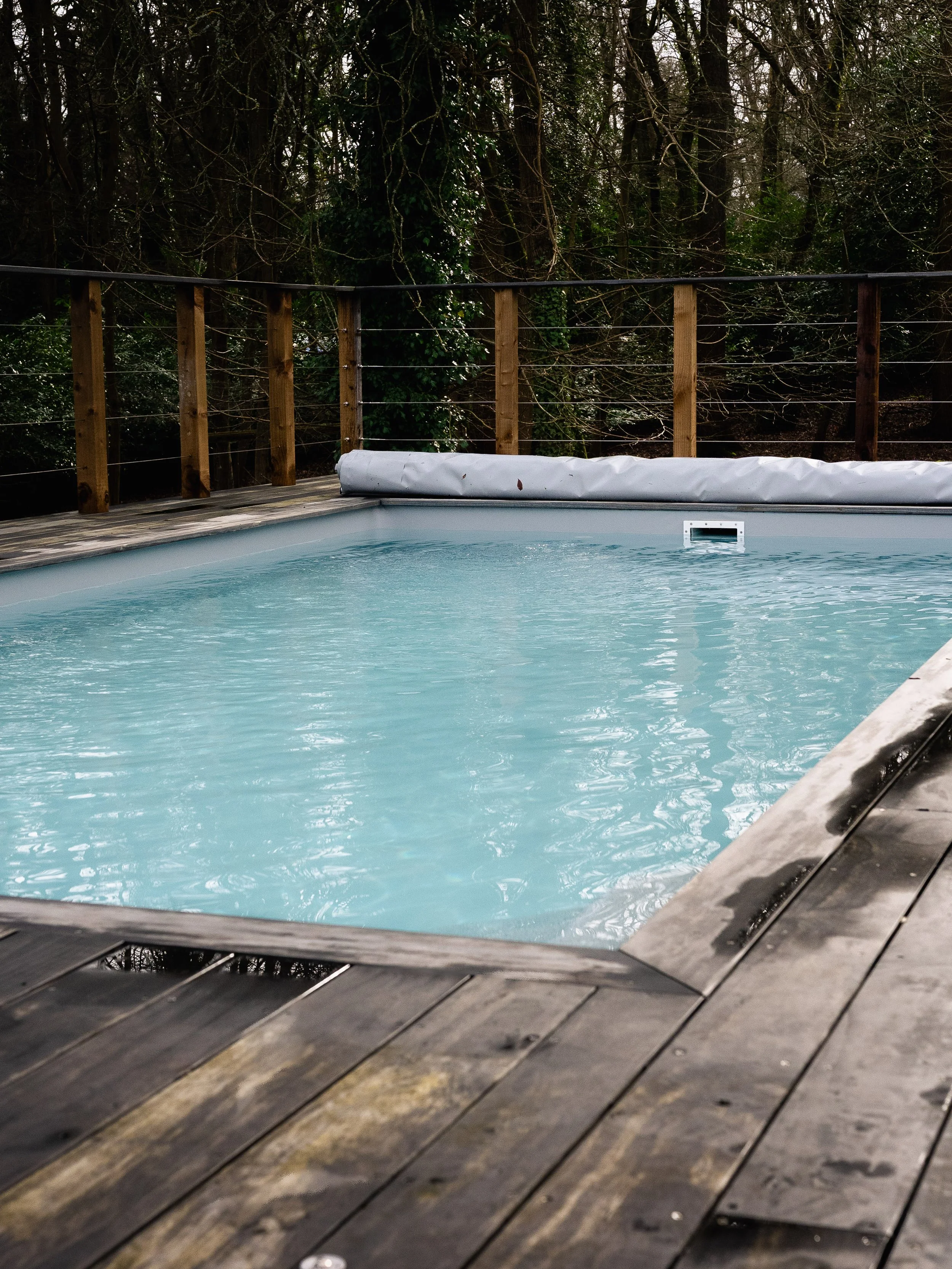 An outdoor swimming pool surrounded by a wooden deck and railing, with trees in the background.