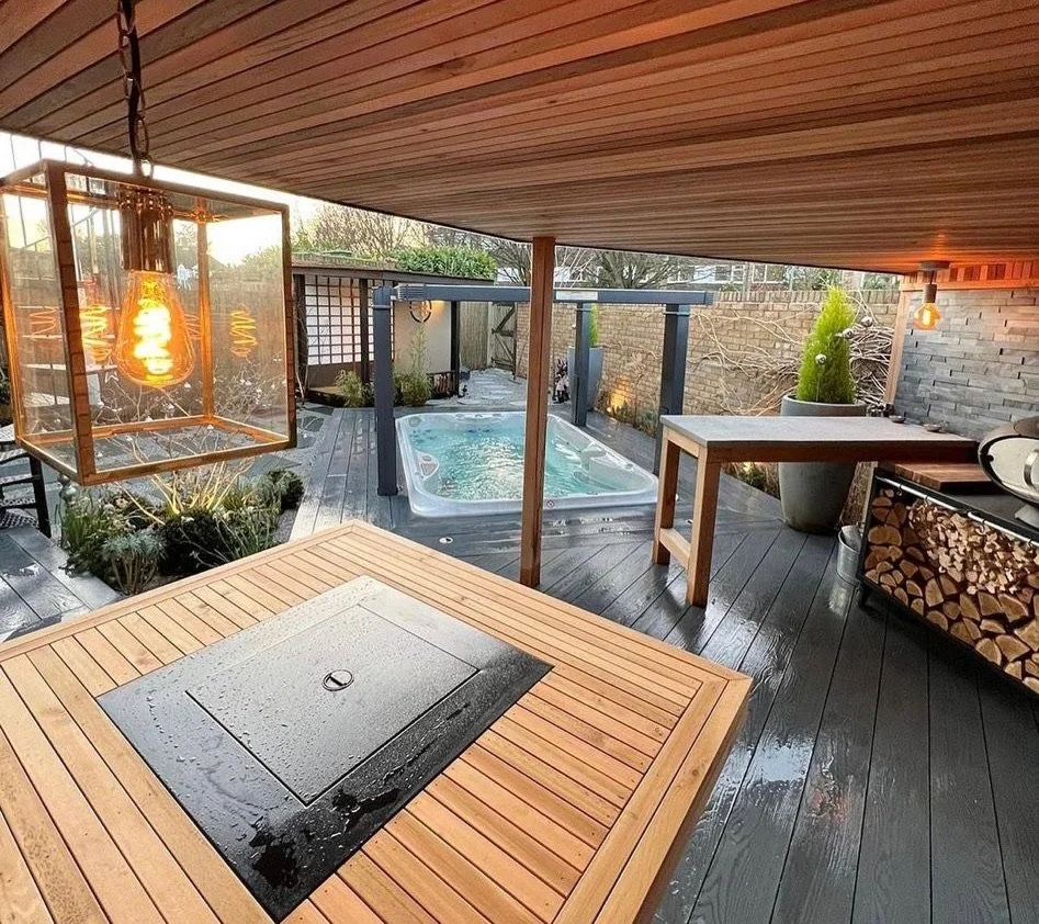 Outdoor spa area with hot tub, wooden deck, and modern design, including a hanging lantern, potted plants, a firepit, and stacked firewood.