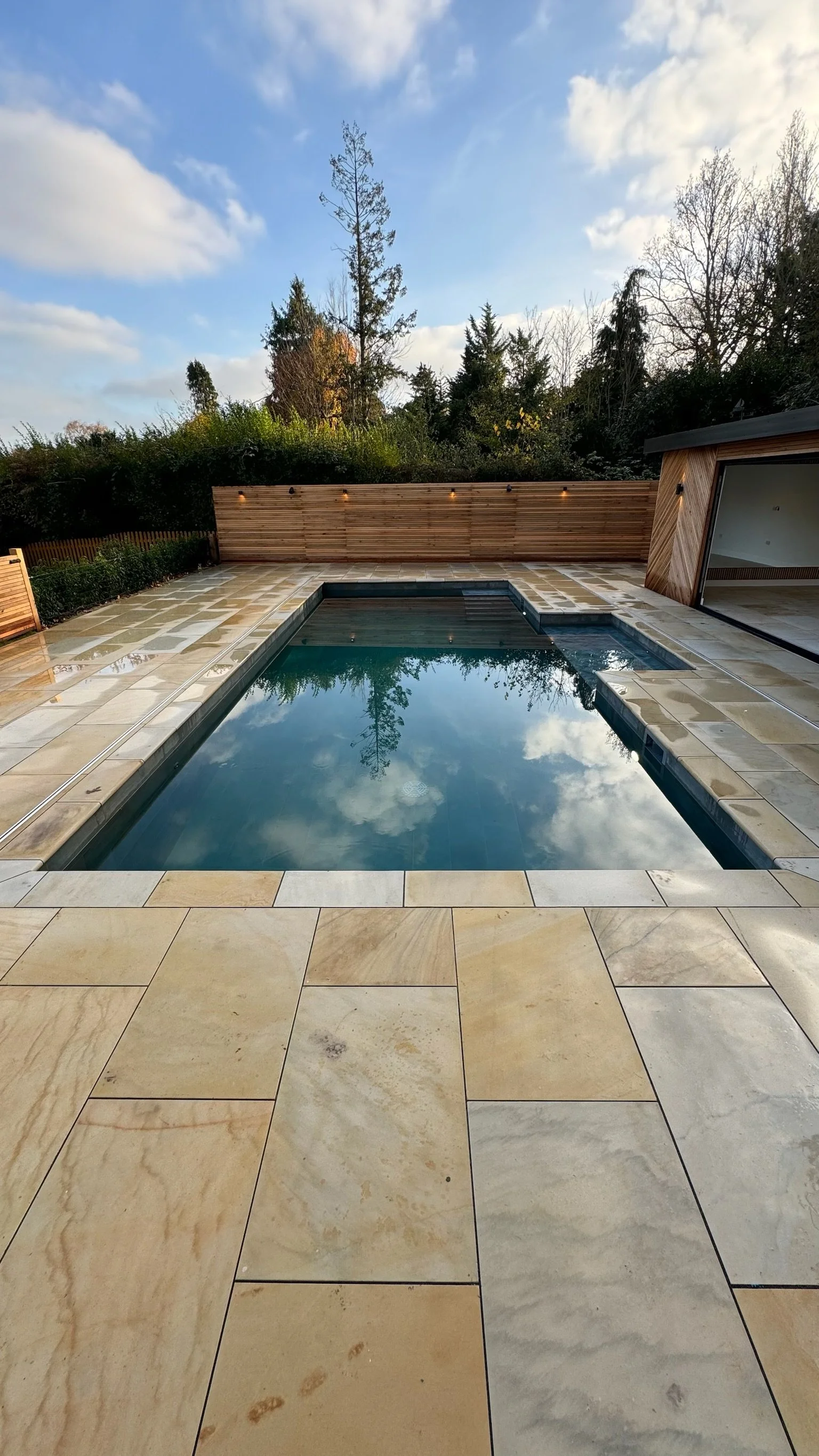 A modern backyard with a rectangular swimming pool reflecting the sky and trees, surrounded by beige stone tiles, with a wooden privacy fence and a small building with sliding glass doors in the background.
