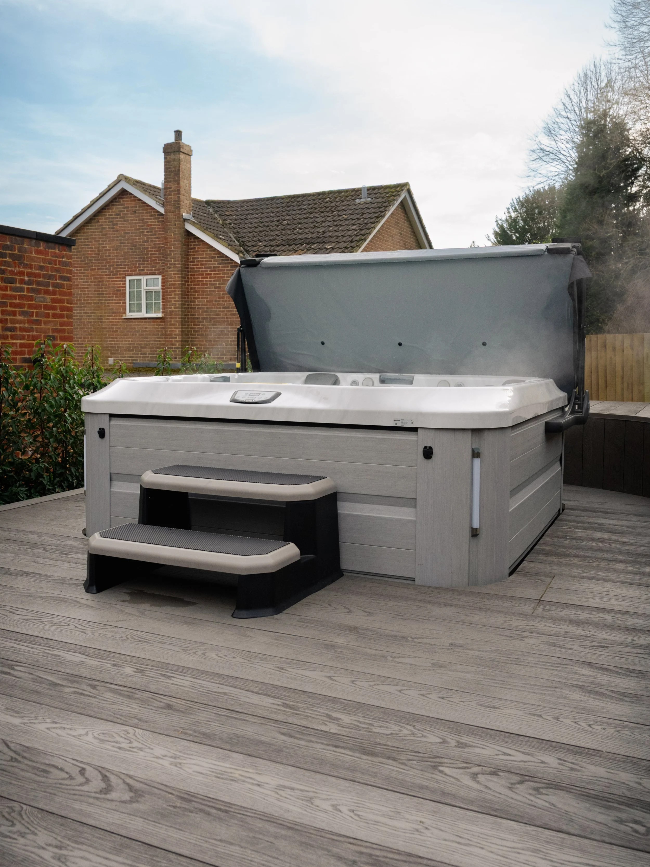 An outdoor hot tub on a wooden deck with steps leading up to it, in a residential backyard with a brick house and a wooden fence in the background.