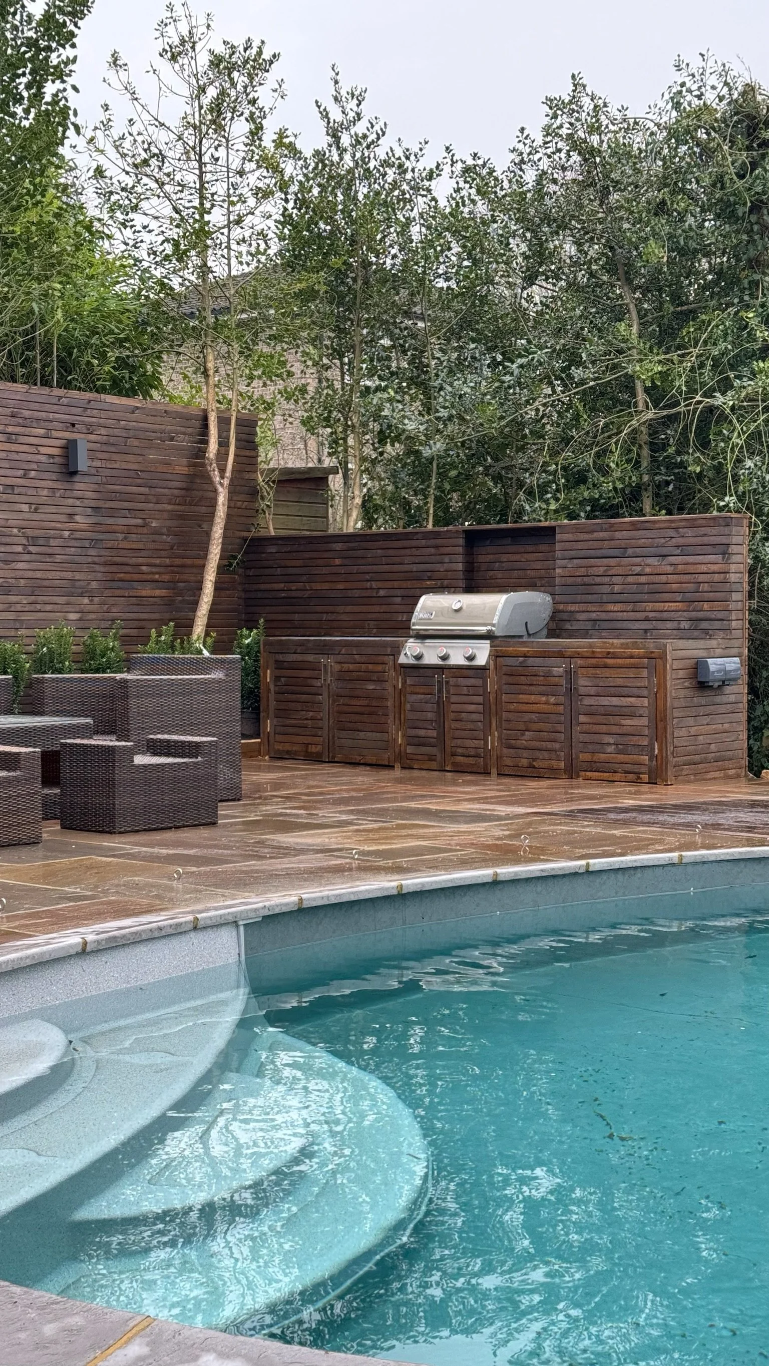 A garden with a swimming pool, outdoor seating, and a wooden outdoor kitchen with trees and greenery surrounding. Expert landscapers and garden designers in Surrey, Hampshire, Berkshire, Camberley, London and surrounding areas.