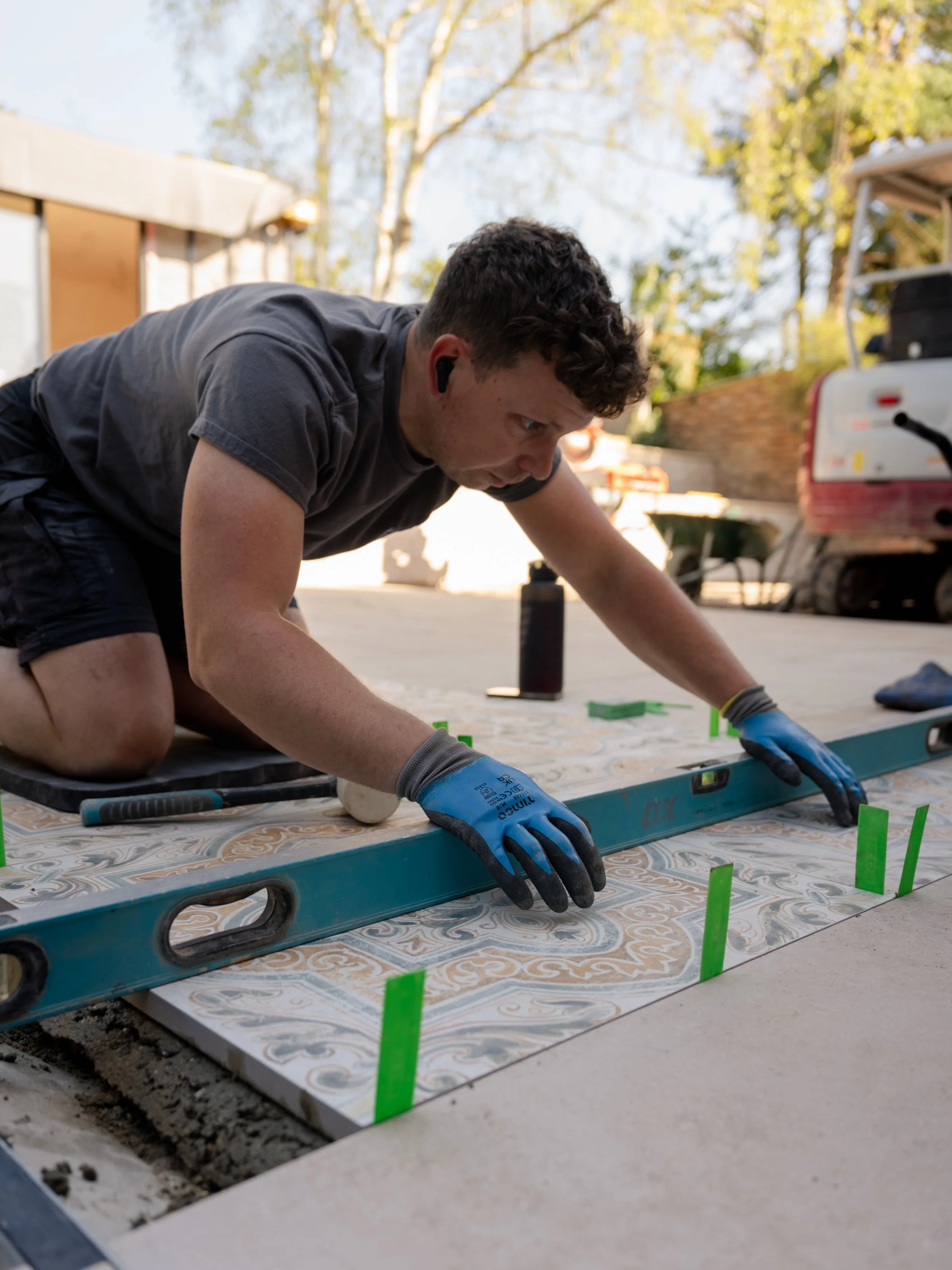 A man kneeling on the ground installing decorative tiles on a surface outdoors. He is wearing gloves and working with tools, with a background of trees and a vehicle.