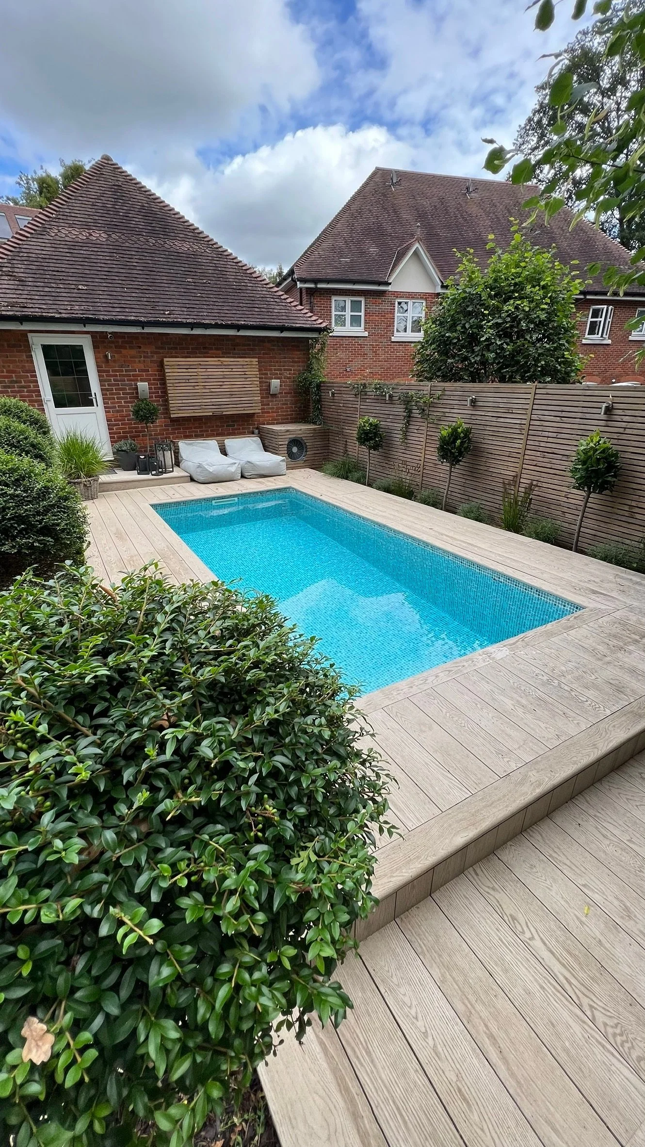 A backyard with a rectangular swimming pool surrounded by a wooden deck, bushes, and small trees, with a brick house and a wooden fence in the background. The sky is partly cloudy.