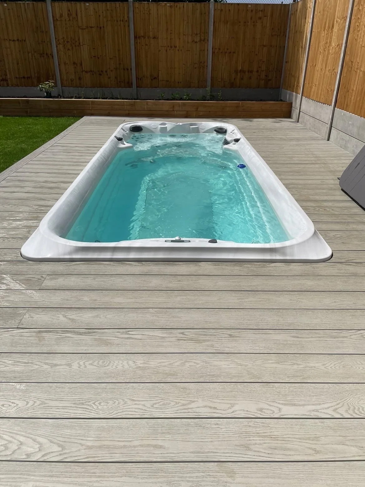 A rectangular hot tub filled with water on a wooden deck in a backyard, with a wooden privacy fence in the background.