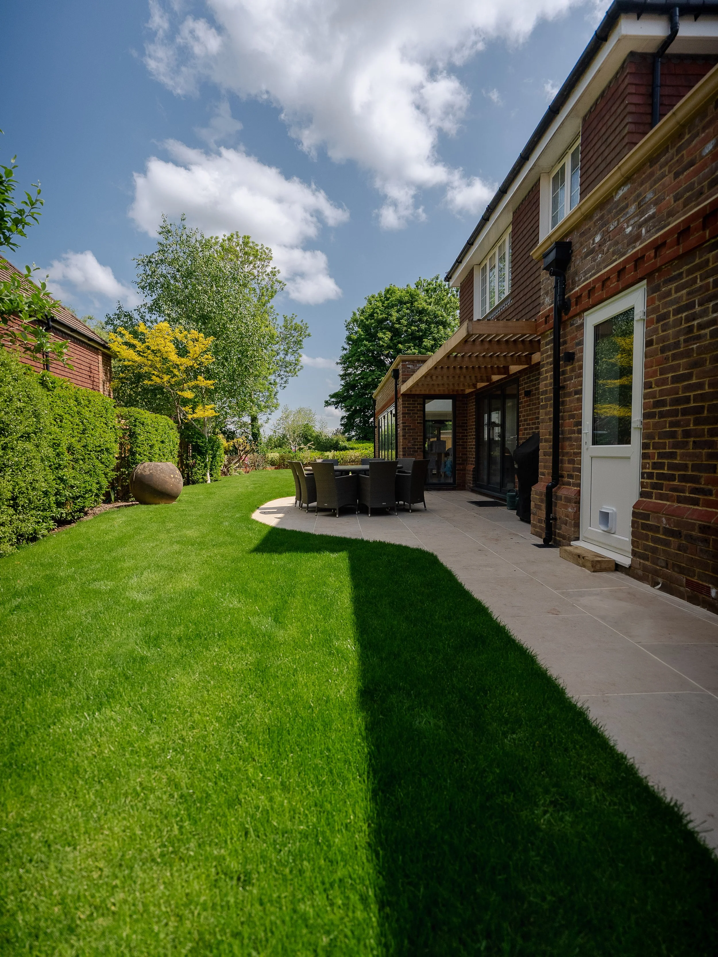 Backyard of a house with a green lawn, patio furniture, bushes, trees, and a partly cloudy sky.