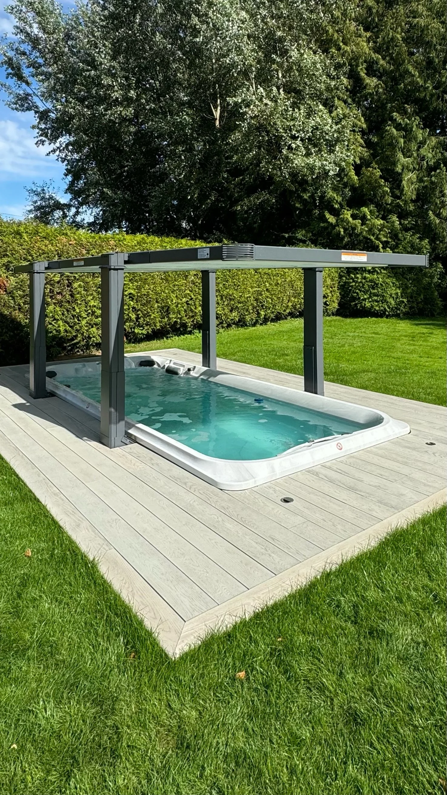 A small outdoor hot tub on a wooden deck with a gray pergola frame, surrounded by green grass and trees.