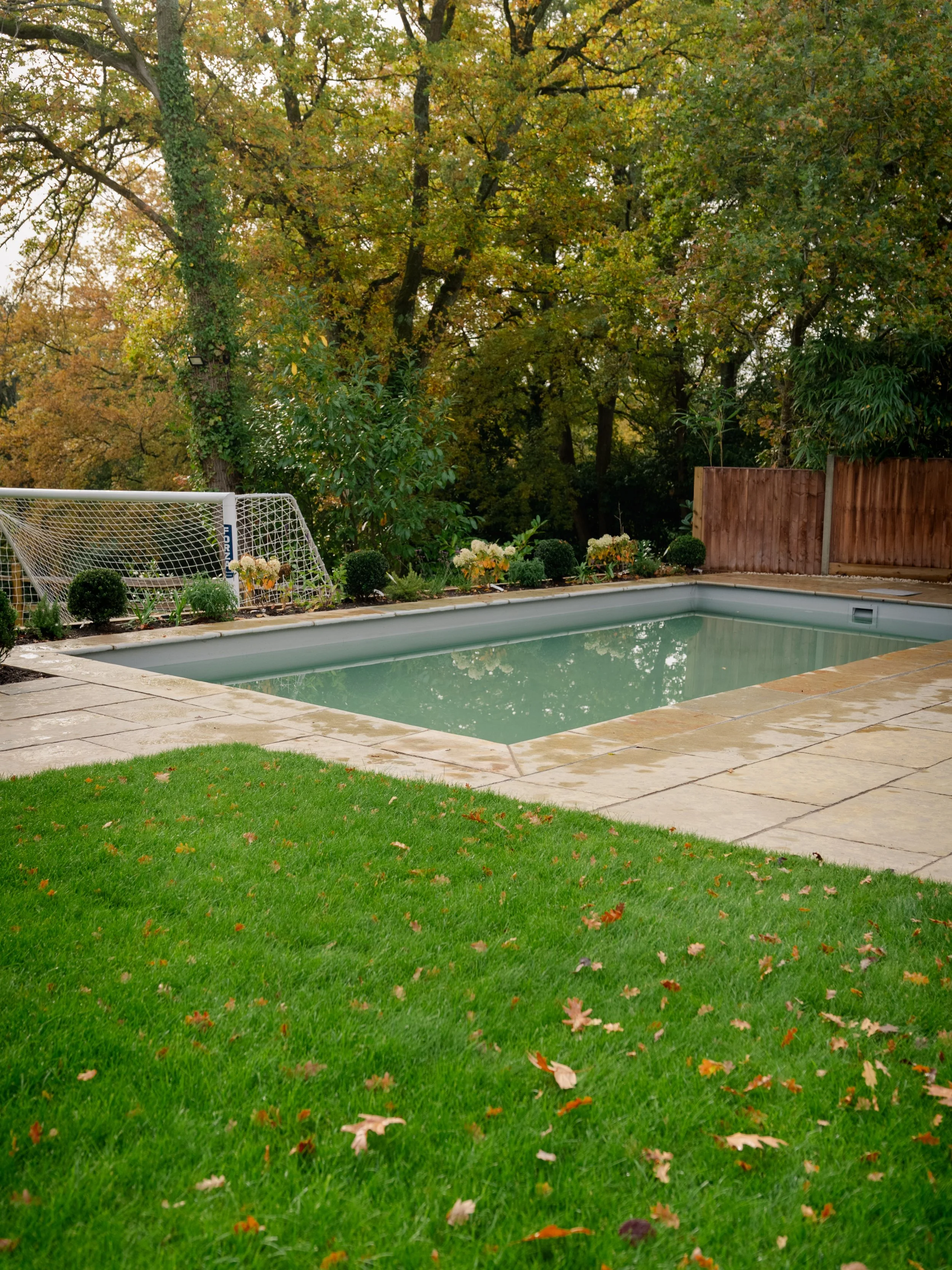 A backyard swimming pool surrounded by a stone deck, with a small soccer goal and potted plants near the fence. The yard has lush green grass and trees with autumn-colored leaves in the background.