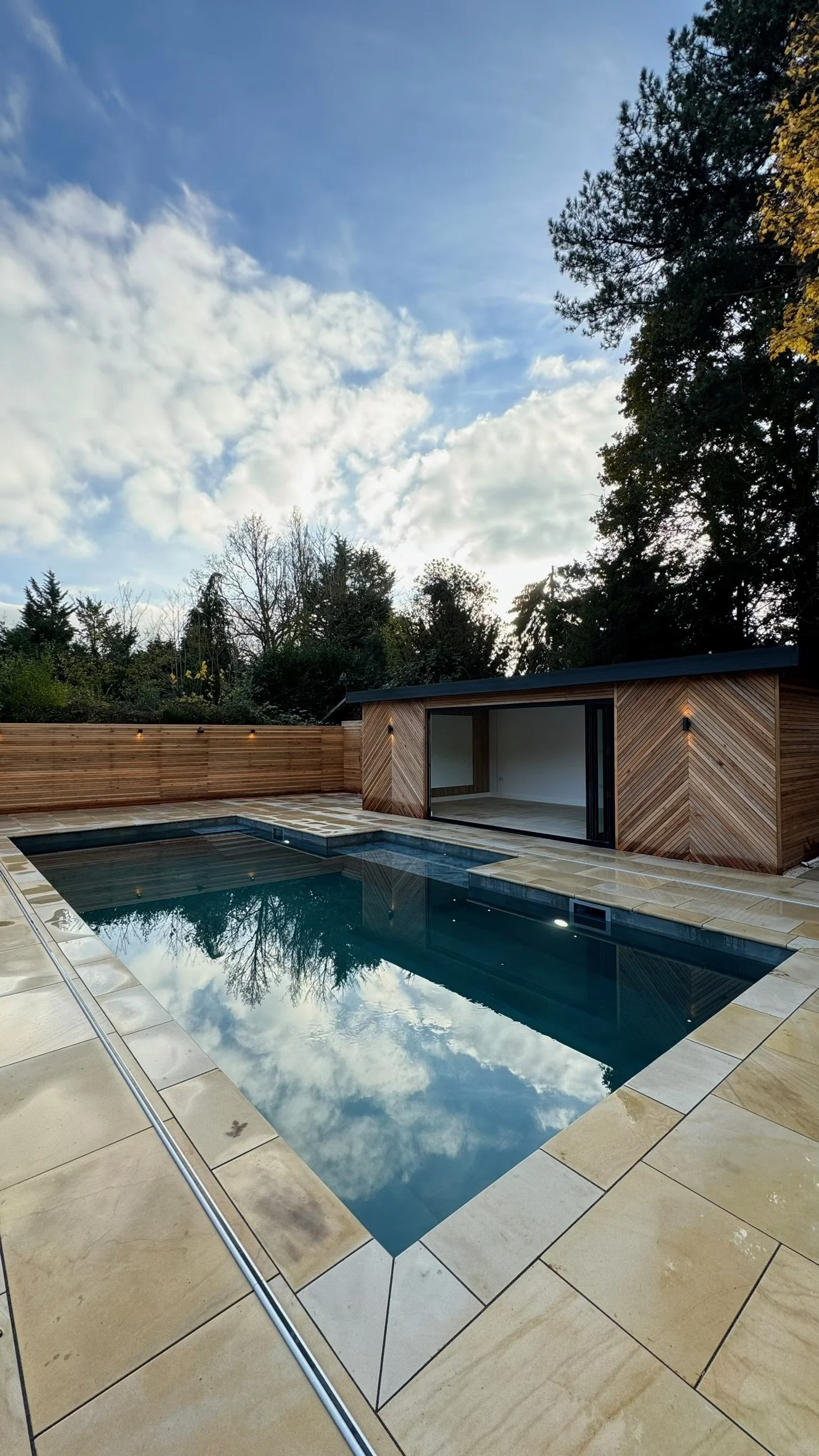 A backyard with a modern swimming pool, surrounded by beige stone paving, and a wooden shed with sliding glass doors, with trees and a cloudy sky in the background.